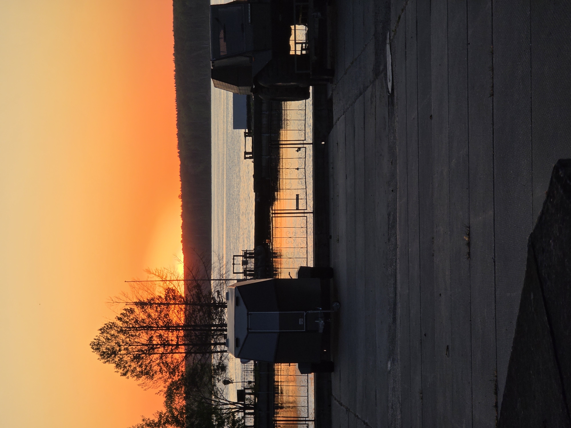 The image shows a sunset scene in Chaikovsky, Russia. A body of water is visible, with reflections of the sunset and some structures. A line of trees is silhouetted against the brightly colored sky. Two dark-colored trailers are parked on a paved surface near a metal fence.