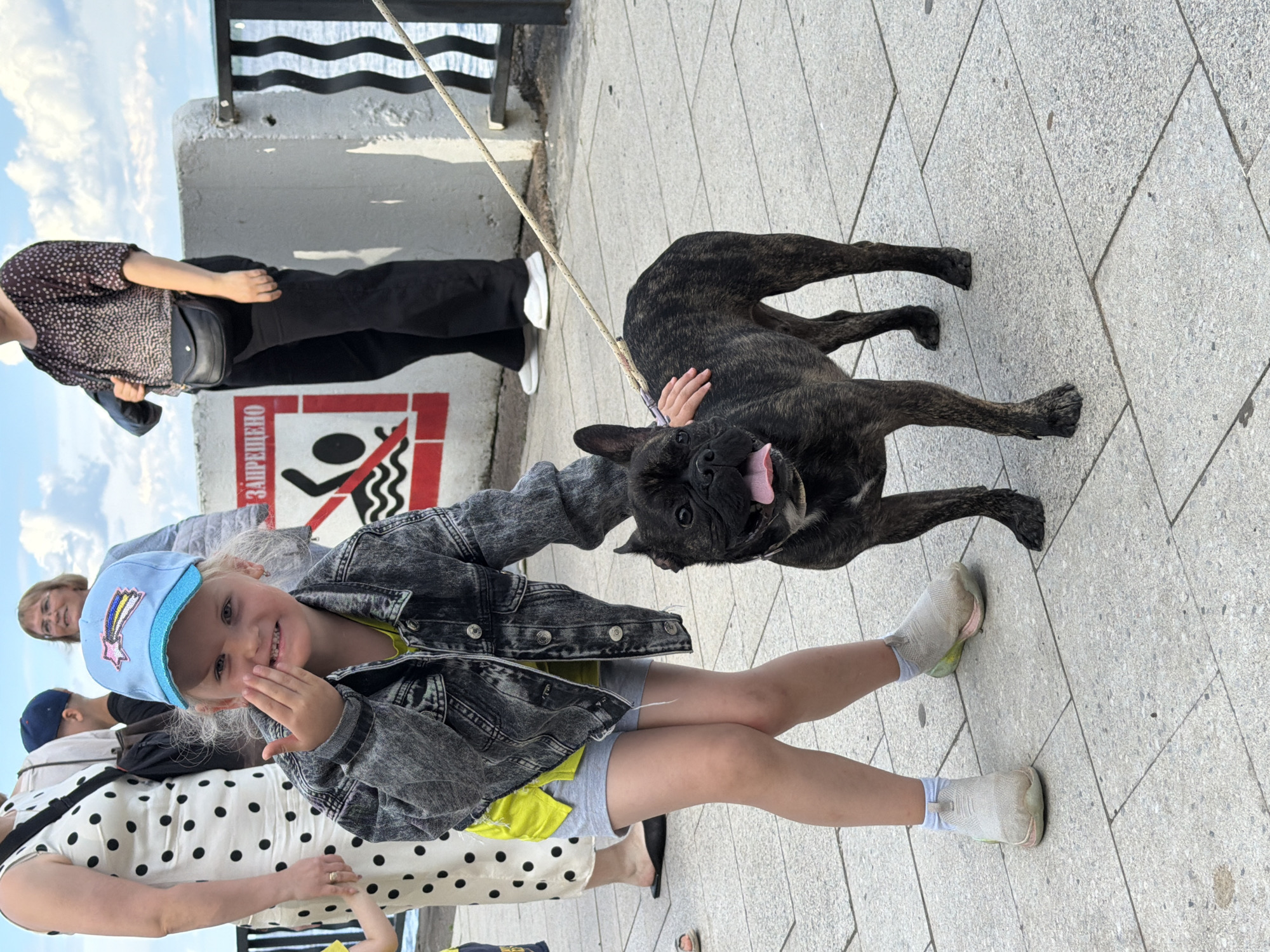 The image shows a girl in a denim jacket and a blue baseball cap with a rainbow and star design. She is smiling and has her hand near her mouth. A brindle French bulldog is standing next to her, with its tongue sticking out. The dog is on a leash held by the girl.
In the background, another person is visible, wearing a patterned top and black pants. There is also a sign with red and white markings that appears to be a warning sign, with a symbol of a person near water and a red circle with a slash through it. The ground is paved with grey tiles.
The scene appears to be outdoors, possibly near a body of water, given the warning sign. The lighting suggests it is daytime. The overall mood is lighthearted and joyful, with the smiling girl and the playful dog.
Visible text: "ЗАПРЕЩЕНО" is visible on the red and white sign, which translates to "FORBIDDEN" in Russian.