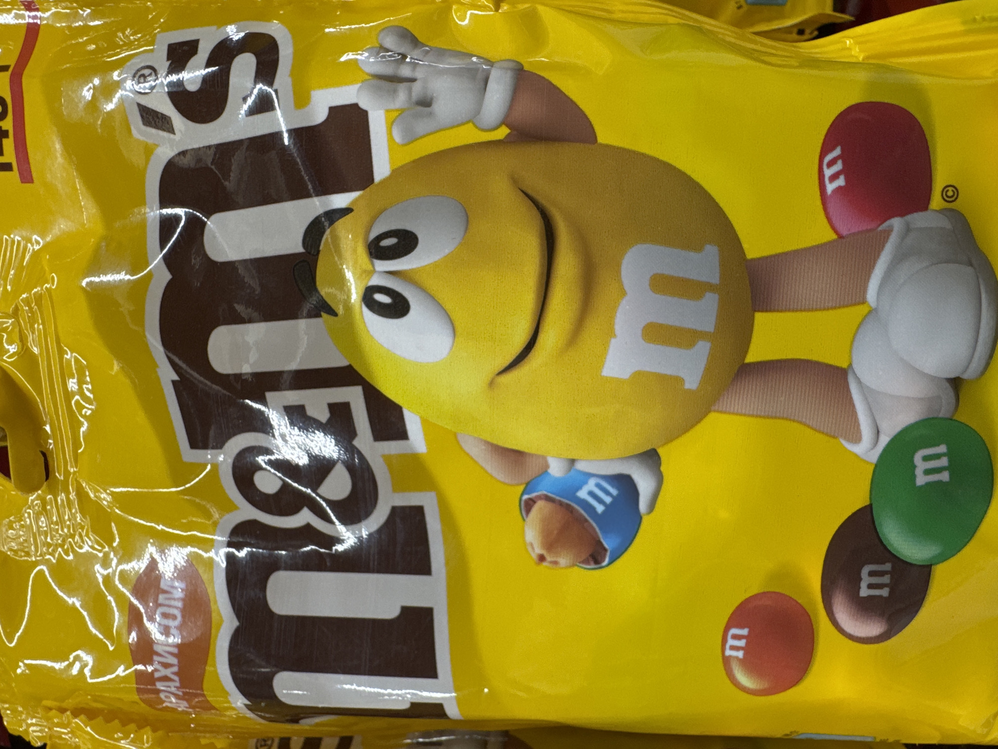 A close-up shot of a yellow M&M's bag with a cartoon of a yellow M&M character on it. The bag contains peanut M&M's, as indicated by the Russian inscription "АРАХИСОМ". The colors of the M&M's candies depicted on the bag are red, green, and brown. No specific location within Ново-Никольское, Russia, or evidence of any interaction with a specific location is seen.