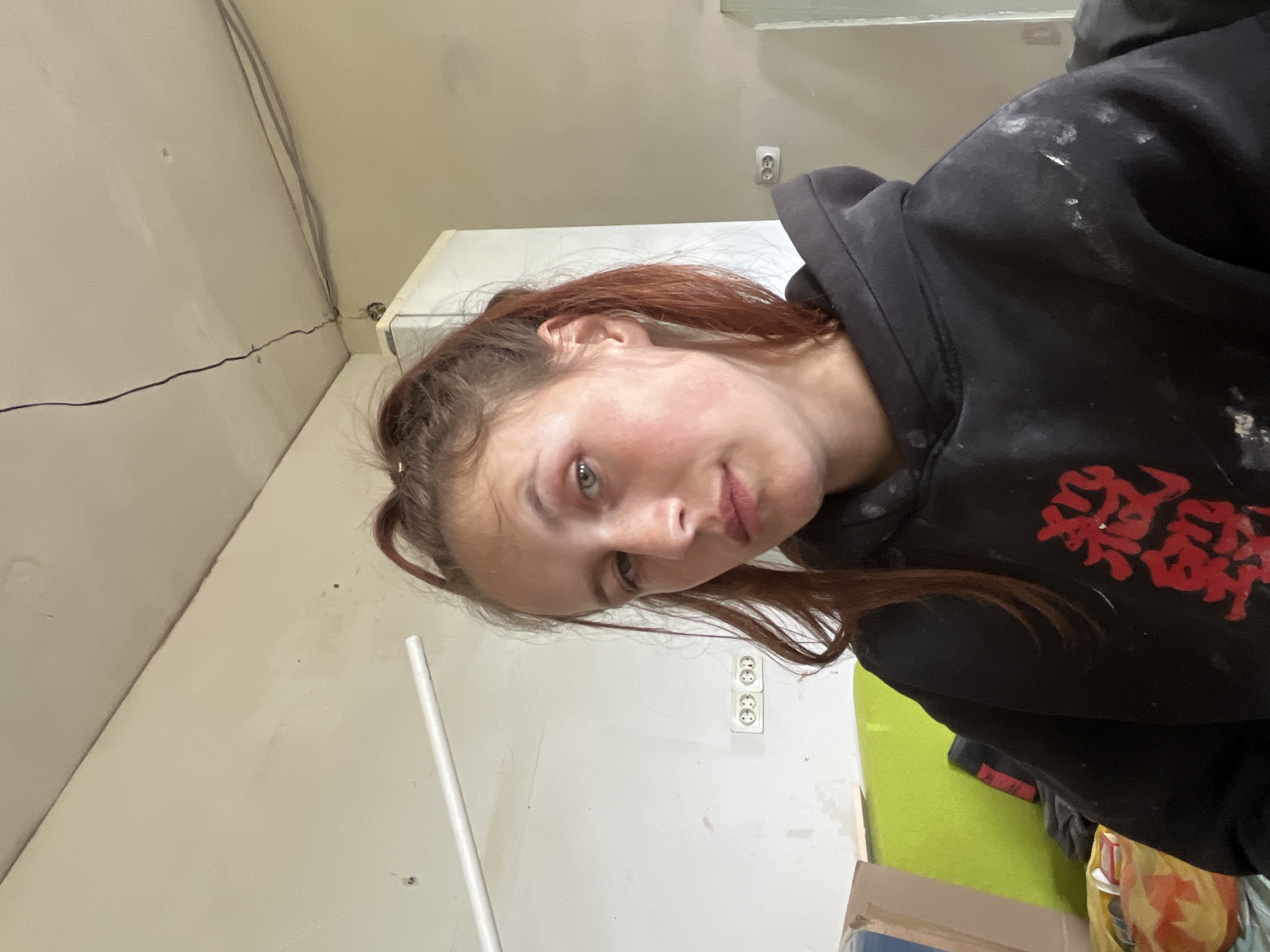 A young person with reddish-brown hair, wearing a black hoodie with red writing on the right shoulder, is facing the camera in an interior setting. The walls are light-colored, possibly cream or light gray, and appear to be under construction or renovation, with exposed wiring visible near the ceiling. There are electrical outlets on the wall behind the person. A white pole leans against the wall on the left. Miscellaneous items and materials, including what appears to be a green surface and cardboard boxes, are partially visible in the lower right corner. The image appears to be taken indoors in Фоки, Russia.