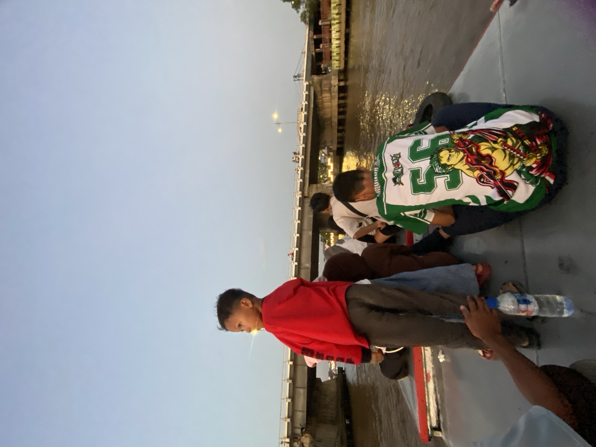This image depicts a boat ride on the Martapura River in Banjarmasin, Indonesia, likely at dusk given the soft lighting and clear sky. A group of people, including several children and adults, are on the boat. In the foreground, a young boy in a red hooded sweatshirt and grey pants stands facing left, looking out at the scenery. Behind him, other passengers are seated. A man in a green and white shirt featuring a character illustration and the number "56" is seated towards the right. The boat is navigating a river with a bridge visible in the background, spanning across the water. Lights from buildings and streetlights illuminate the riverbanks and the bridge. The water is calm, reflecting the ambient light.