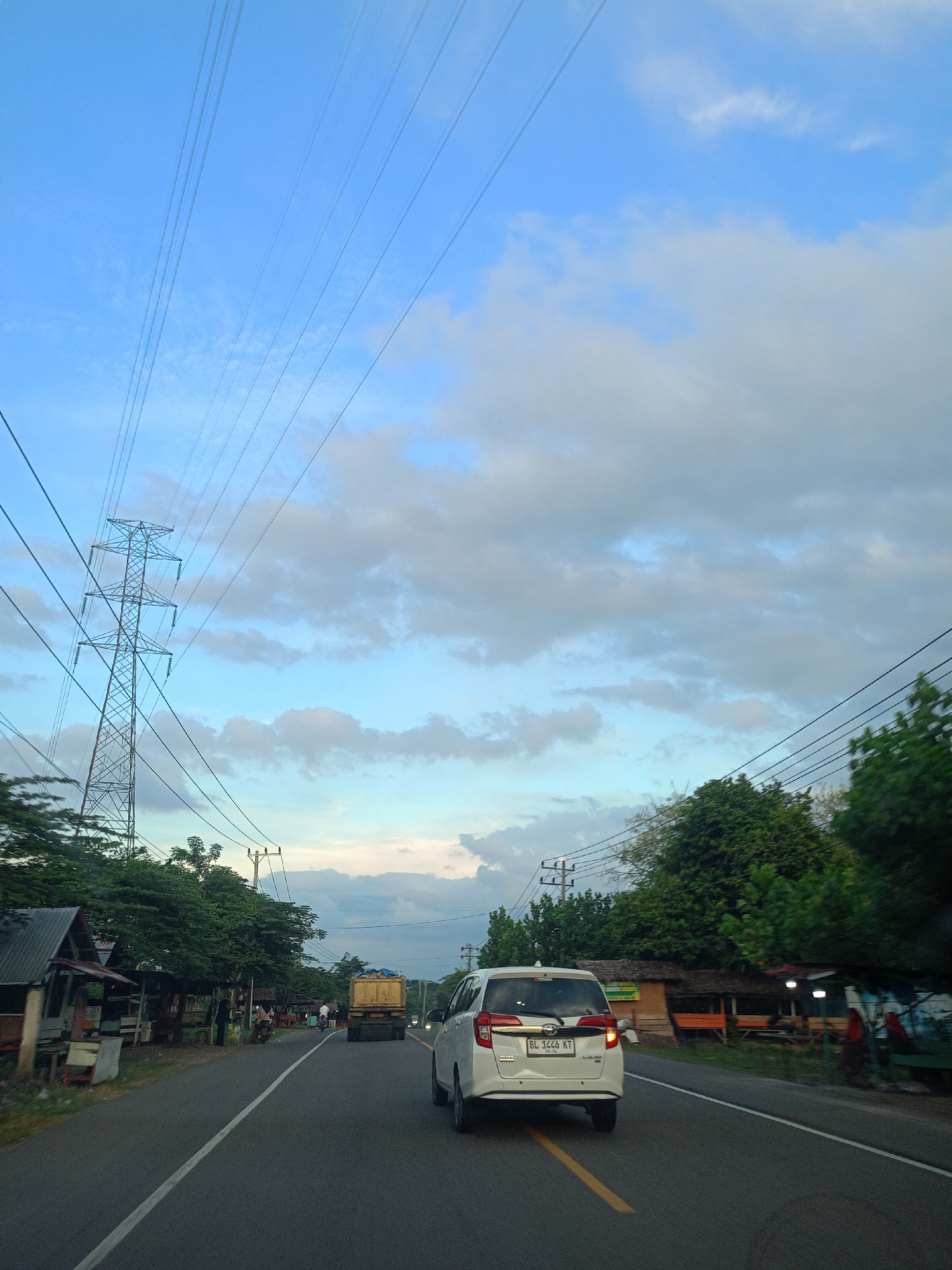 The image depicts a road in Lhokdagang, Indonesia. A white car with license plate "BL 1446 KT" is traveling on the right lane. A yellow truck is visible further down the road, as well as other cars. A motorcycle with two passengers is visible further down the left lane. On either side of the road are low buildings, small shops, and trees. An electrical transmission tower is visible on the left side of the road. The sky is blue with patches of white clouds. Overhead electrical lines stretch across the sky. The road has a solid white line marking the left side of the lane. The weather appears to be clear and sunny.