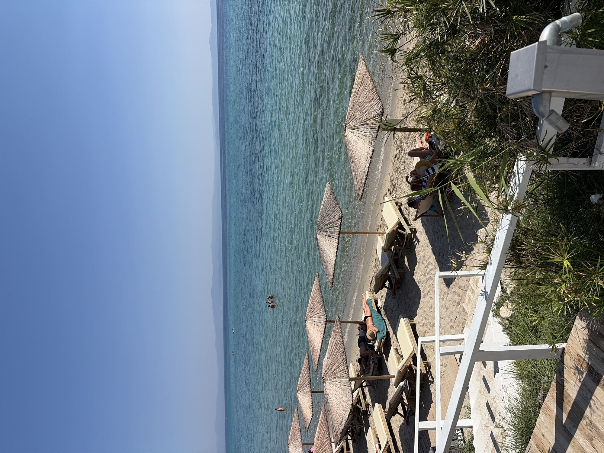 A bright, sunny day scene depicts a tranquil beach in Kassandra Municipal Unit, Greece. The foreground features a wooden pathway leading to white stairs bordered by lush green vegetation. Several empty sun loungers are arranged on the sandy shore, some under thatched umbrellas. In the middle ground, a few people are visible relaxing on loungers, one woman wearing a sun hat. The crystal-clear turquoise water of the Aegean Sea stretches to the horizon, where faint hills are visible. Several small boats are scattered across the water, and a few individuals are swimming near the shore. The scene conveys a peaceful atmosphere characteristic of a Mediterranean beach vacation.