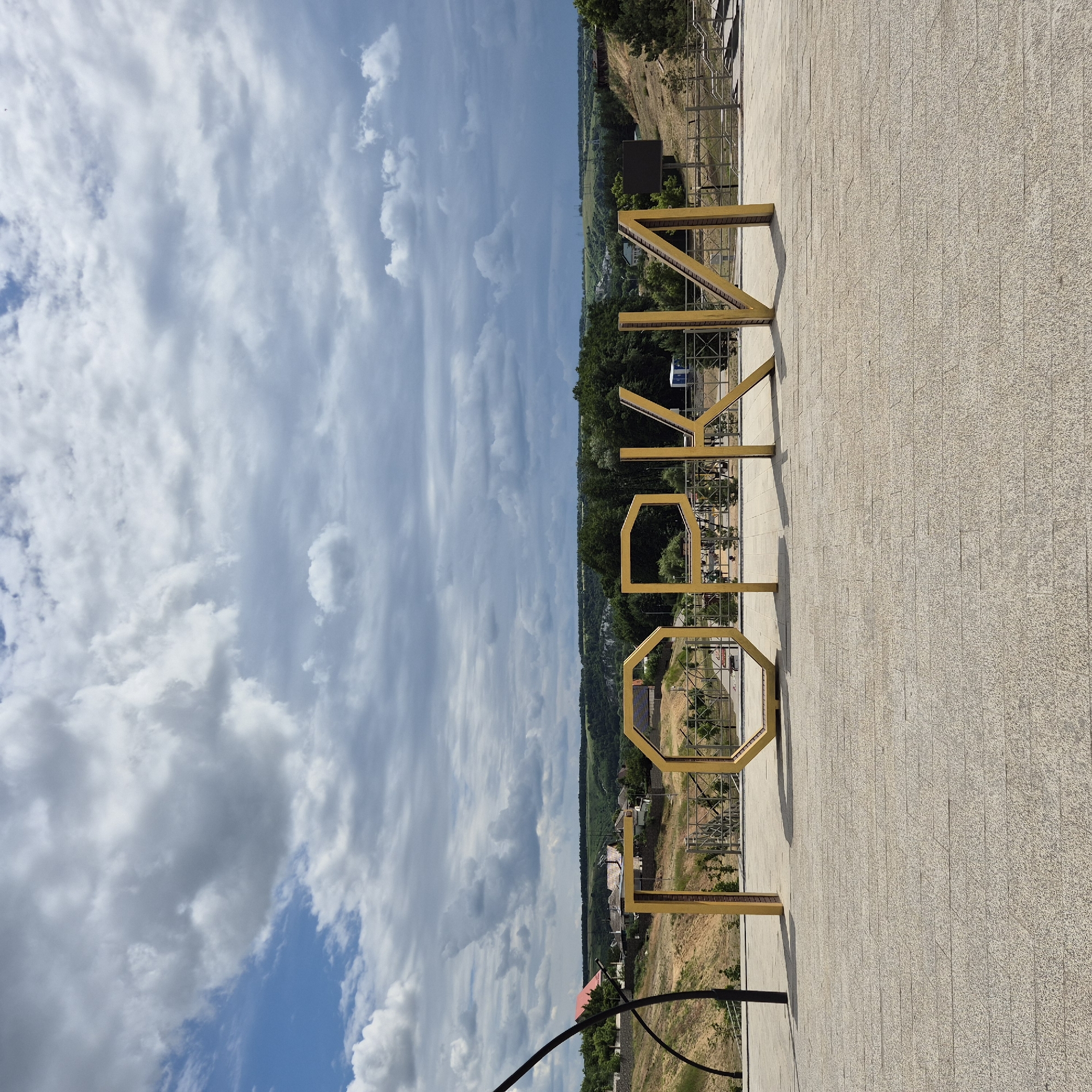 In the city of Liski, Russia, an outdoor scene features large, golden, block-style letters spelling out "ГОРКИ" (GORKI) in the foreground. The letters are positioned on a light-colored, textured stone pavement. Behind the letters, a landscaped area with a retaining wall and some vegetation is visible, leading up to a hillside. Further in the background, a small town or village with houses and trees can be seen nestled in a valley. The sky above is filled with dramatic, grey and white clouds against a blue sky. The overall impression is of an urban park or recreational area.