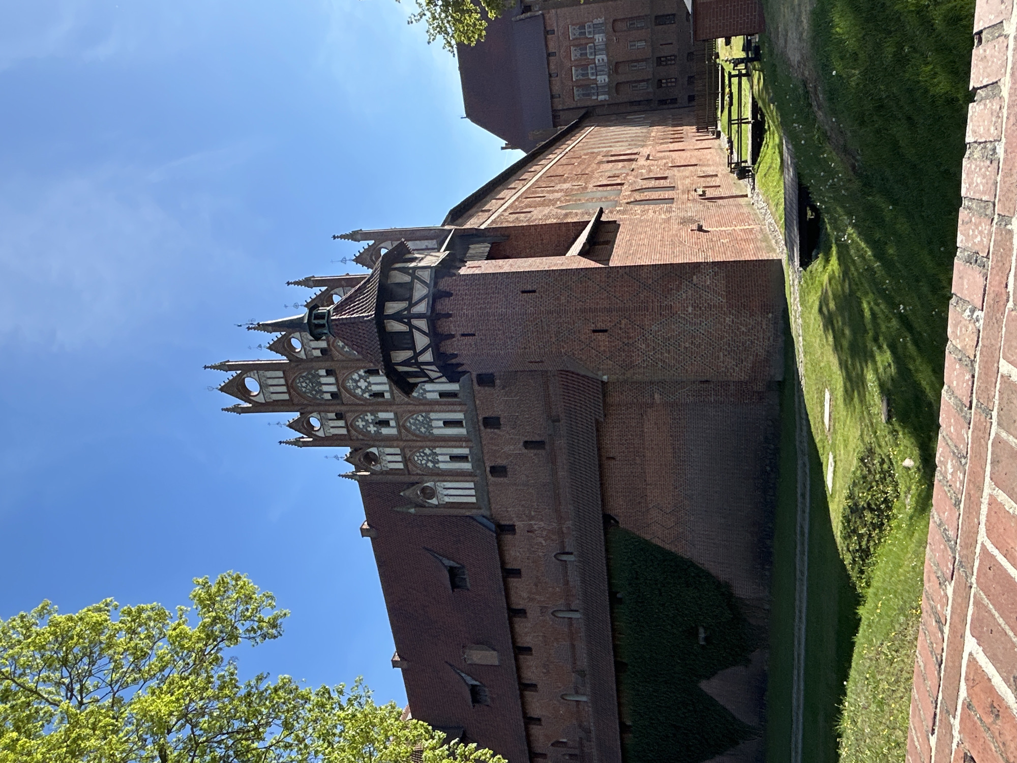 The image shows a view of Malbork Castle in Malbork, Poland. The castle is made of red brick and features Gothic architecture. A tall, decorated tower with pointed arches and a red-tiled roof is visible. Adjacent to the tower, there's a section with half-timbered detailing. A long, brick wall runs alongside a grassy moat or ditch. The sky is blue. Green trees are also visible.
