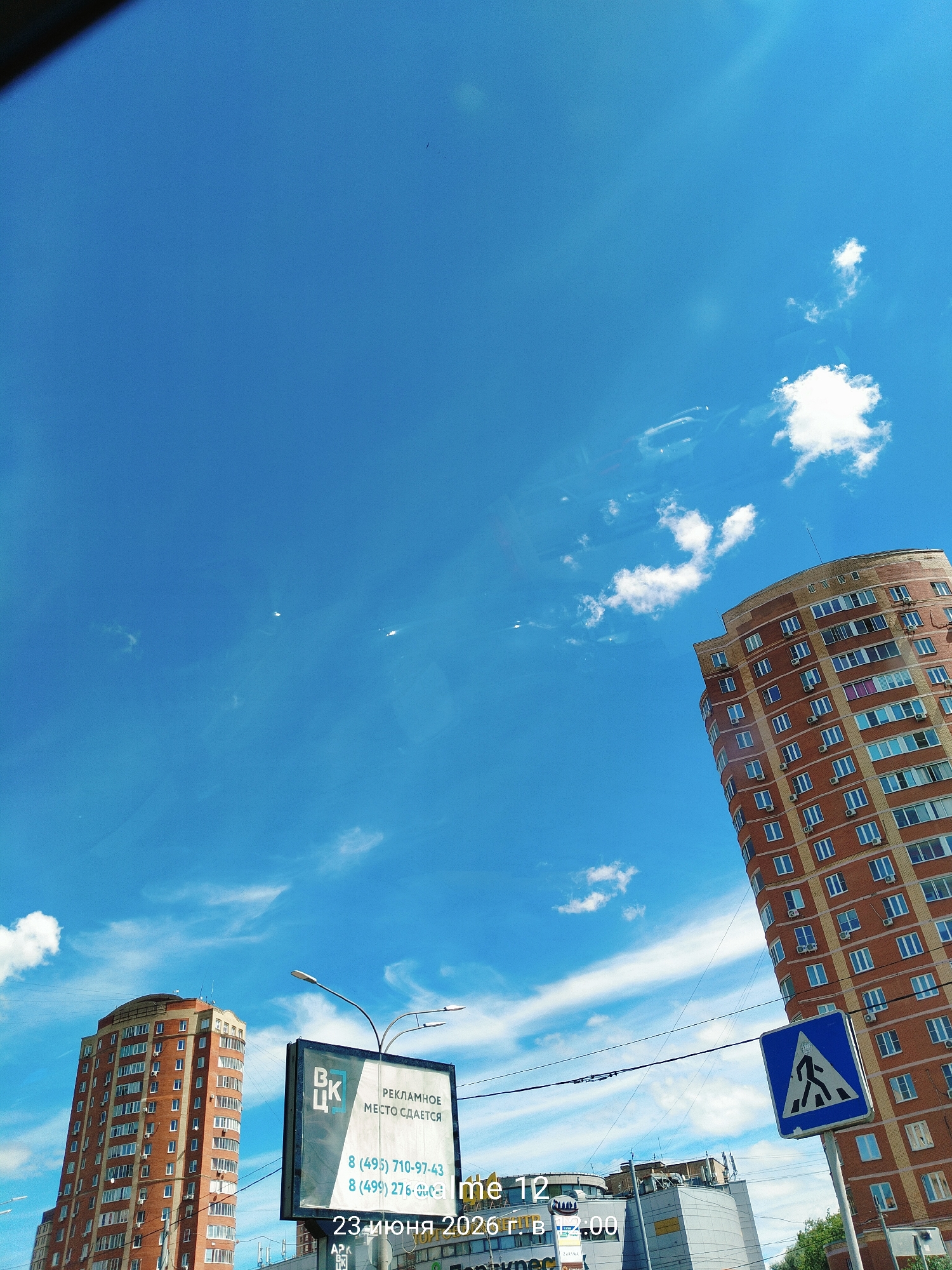 This media file captures an urban scene under a bright blue sky with scattered clouds, suggesting a sunny day, likely midday due to the clear sky and the presence of the sun's reflection on surfaces. The primary subjects are two tall, modern residential buildings with red brick facades and numerous windows. The building on the right is more prominently featured, curving slightly and dominating the right side of the frame.
In the foreground, a billboard advertises "РЕКЛАМНОЕ МЕСТО СДАЕТСЯ" (Advertising space for rent) with contact phone numbers: 8 (495) 710-97-43 and 8 (499) 276. Below this, text indicates "23 июня 2026 г. в 12:00" (June 23, 2026, at 12:00), suggesting the image might be a screenshot or a digital composite with a timestamp. A blue pedestrian crossing sign with a white silhouette of a person walking is visible on a pole, indicating a street or intersection.
In the background, the top of another building with a curved, modern architectural style is visible, along with some greenery, suggesting a developed urban or suburban area. The overall impression is one of a typical day in a city, with advertisements, infrastructure, and residential buildings under a clear, expansive sky. The presence of what appears to be reflections on the surface where the photo was taken adds a layered, almost ethereal quality to the scene.