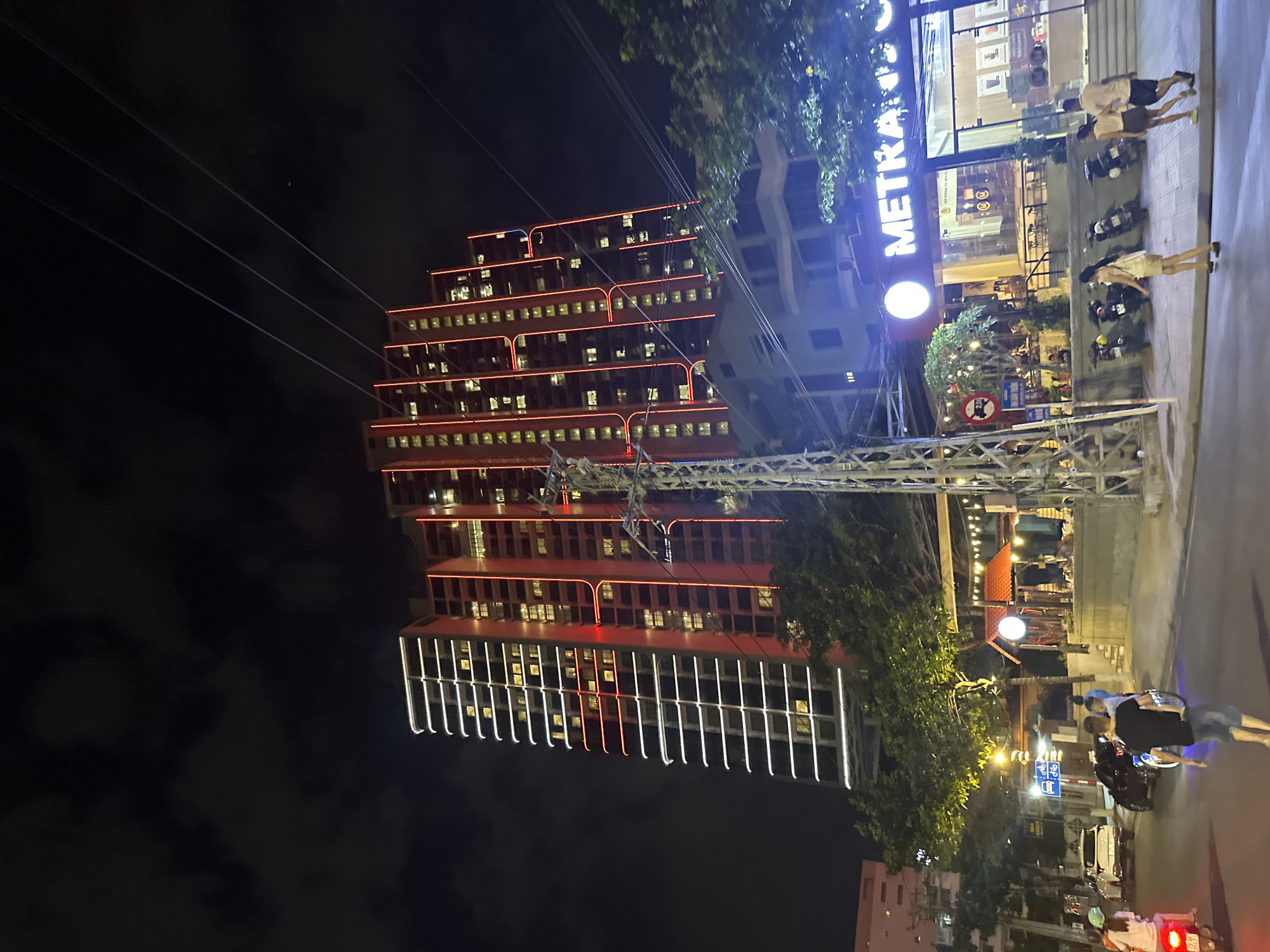 The scene at night in Bac Nha Trang Ward, Vietnam, includes a high-rise building illuminated with both red and white neon lights, and a Metra coffee shop nearby. People are walking and riding scooters on the street. Streetlights illuminate the area. Overhead wires are visible against the dark sky.