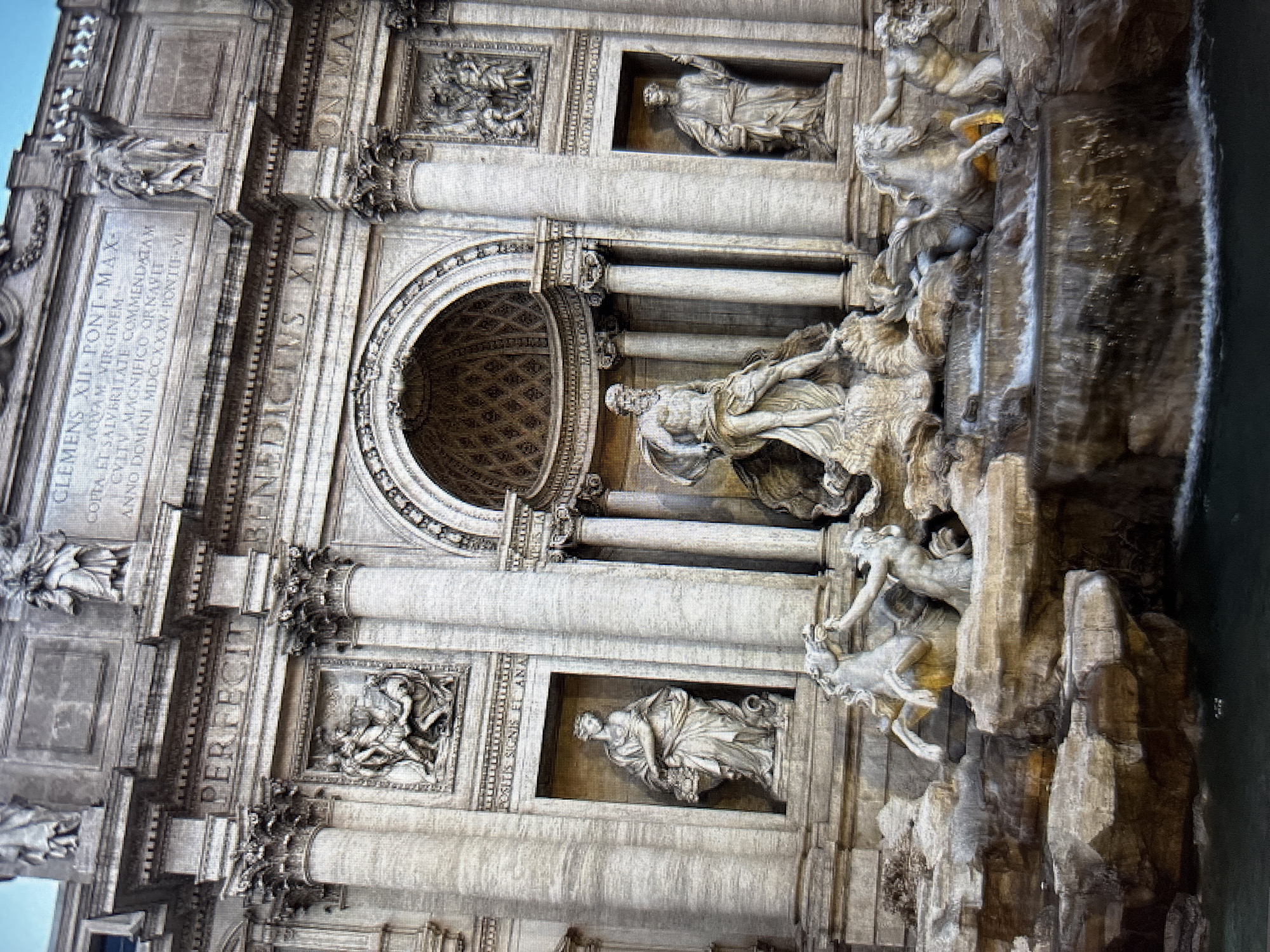 The image shows a close-up of the Trevi Fountain in Rome, Italy. The fountain is a large Baroque fountain featuring a central figure of Oceanus being pulled by a chariot driven by two Tritons, with horses and other mythical creatures. The fountain is made of travertine limestone and is adorned with numerous statues and sculptures, including allegorical figures representing fertility and abundance. The water cascades down rocky formations into a large basin. The architecture surrounding the fountain is ornate, with columns, pediments, and niches containing statues. The lighting suggests it is daytime, and the sky is visible in the upper portion of the image. The fountain is a popular tourist attraction, and the image captures its grandeur and intricate details.