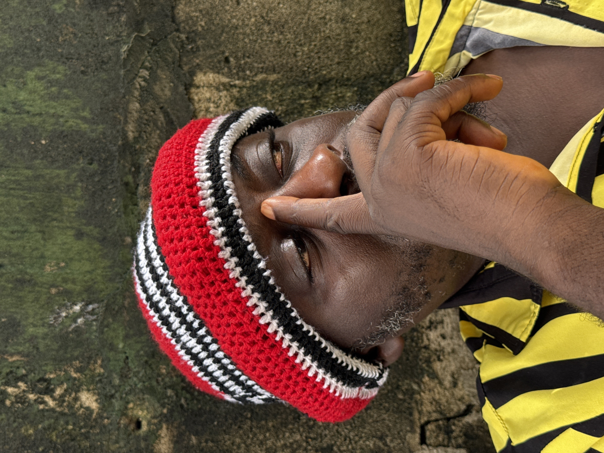 A man wearing a red, white, and black striped beanie is lying on what appears to be a weathered concrete surface. He is wearing a yellow and black striped shirt. His hand is near his face, touching his nose. The background suggests an outdoor environment in Ado, Nigeria, perhaps a weathered wall or ground.