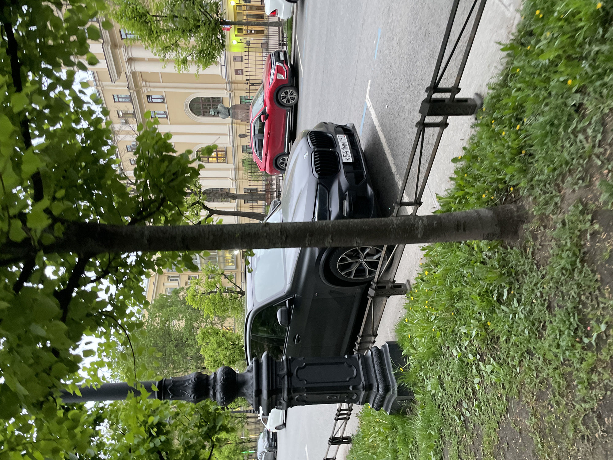The image shows a street scene in Saint Petersburg, Russia. A black BMW X6 is parked on the street next to a red SUV. A sidewalk is partially visible, with a metal fence separating it from the street. Trees and greenery are present, partially obscuring the view of a building in the background. The building appears to be a historical structure. A lamppost is visible in the foreground. The license plate of the BMW is visible.