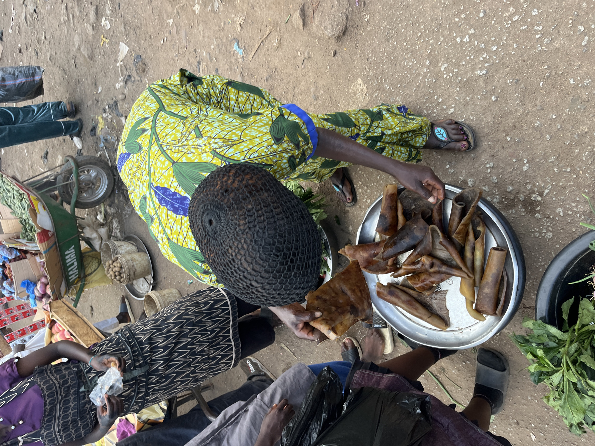 In Bukuru, Nigeria, a person wearing a vibrant yellow and green patterned dress and a black woven hairstyle is bending over a metal tray filled with dried meat. Their hands are occupied, one holding a piece of the meat while the other appears to be arranging or selecting from the tray. The ground is dusty and appears to be a market setting, with a wheelbarrow and other goods visible in the background, hinting at a bustling commercial environment. The overall scene captures a moment of transaction or preparation of food in a Nigerian market.