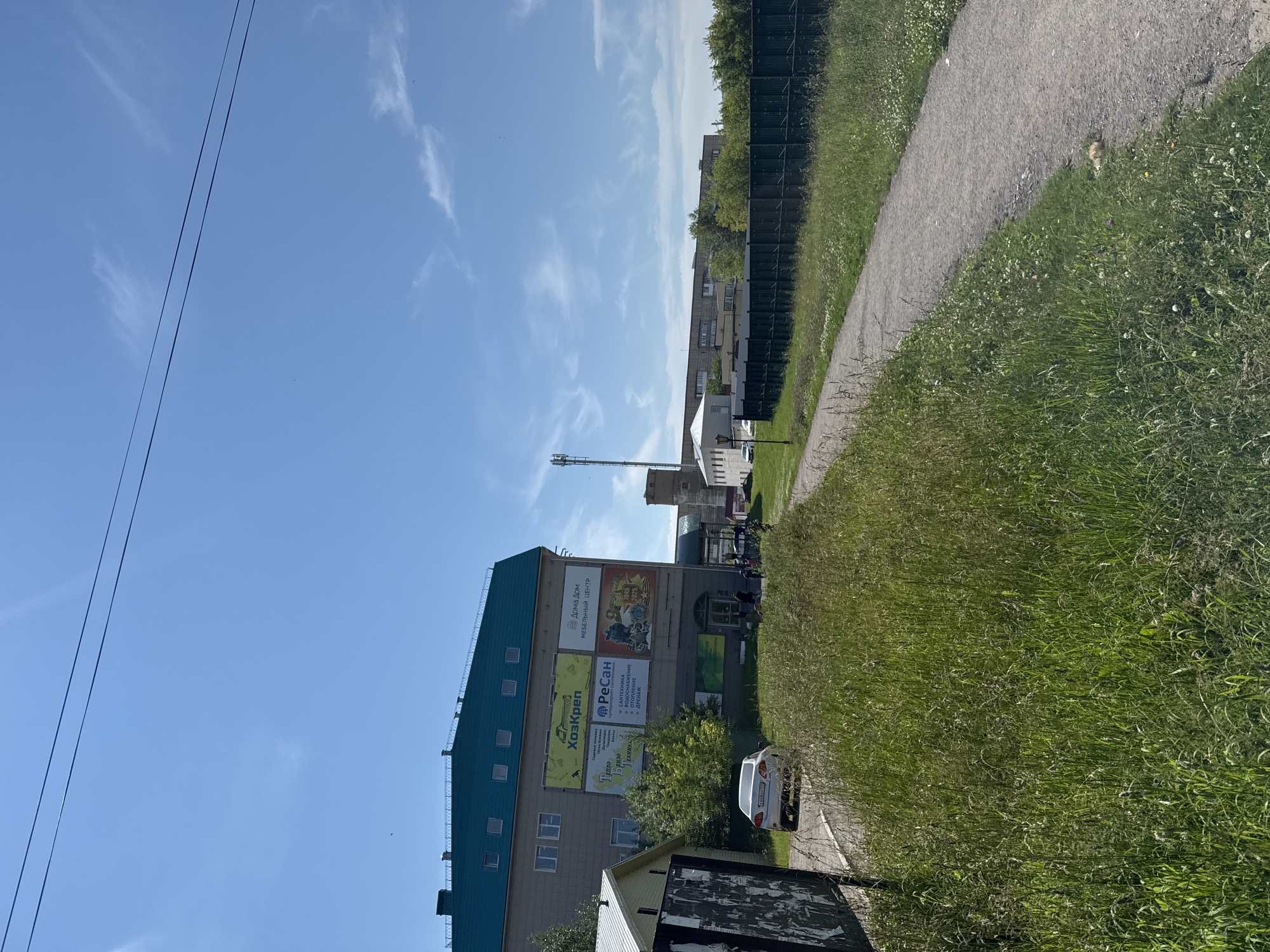 The image depicts an outdoor scene on a sunny day in Vereshchagino, Russia. In the foreground, a gravel pathway winds through tall, green grass dotted with small white flowers. To the left, a white car is parked on a paved area beside a low building with a white roof. Further into the scene, a multi-story building with a green roof and several advertisements on its facade is visible. One advertisement features Russian text and images, including a yellow logo with Cyrillic lettering that reads "Хоскрен". Another sign has the text "Дом Дом Мебельный Центр". Behind this building, a tall cell tower stands prominently against the blue sky, with scattered white clouds. In the background, other buildings and trees are discernible, along with a dark metal fence. A few people are walking near the entrance of the multi-story building. The overall environment suggests a suburban or semi-urban setting.