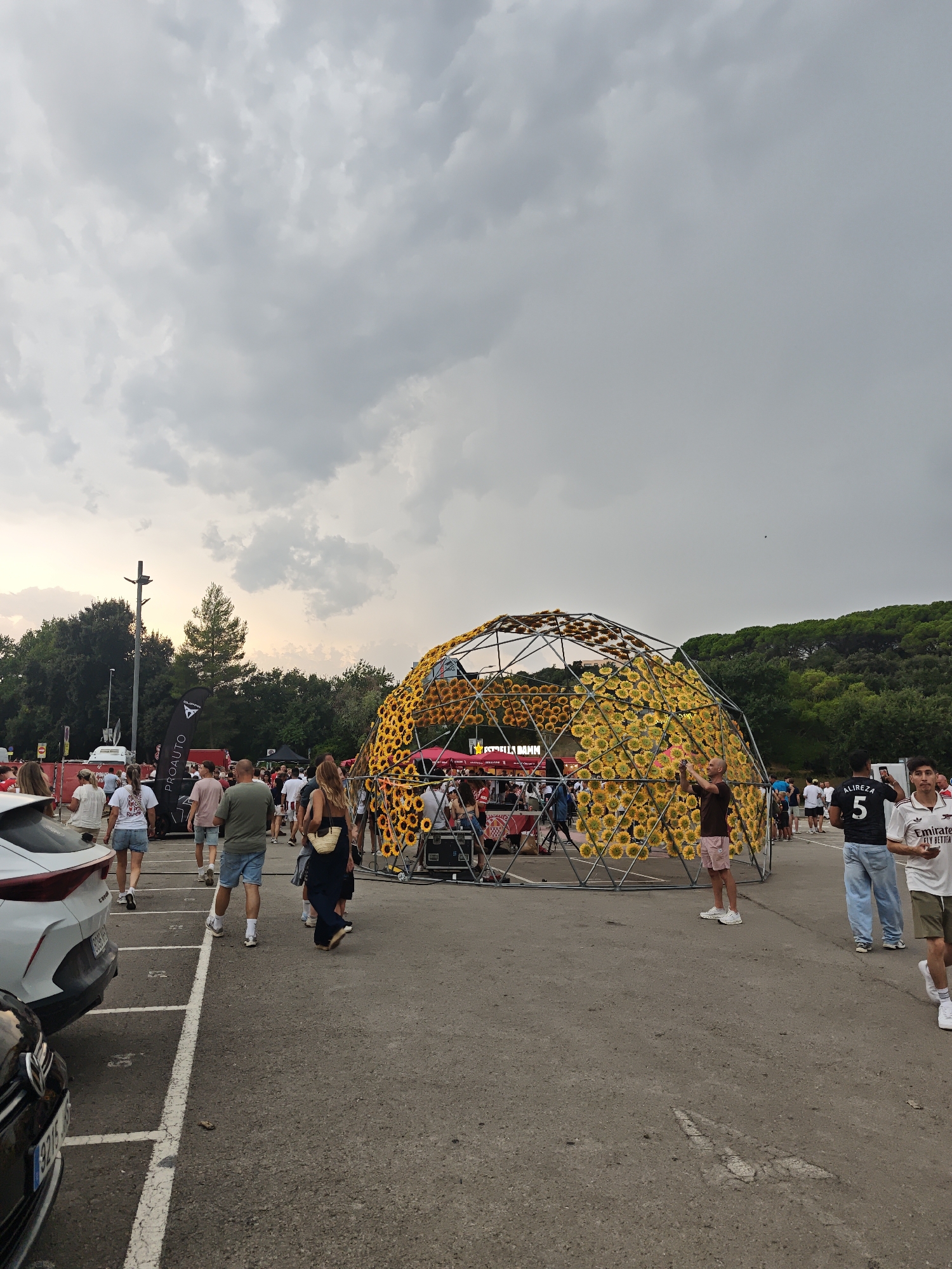 The image captures an outdoor gathering with a geodesic dome structure adorned with sunflowers as the focal point. A crowd of people mills about the paved area, which appears to be a parking lot. The sky is overcast with dramatic clouds. To the left, cars are parked, including a white SUV and a black sedan. In the mid-ground, a woman with blonde hair walks away from the camera, carrying a woven tote bag. To her right, a man wearing a black t-shirt with the name "ALIREZA" and the number "5" printed on the back stands facing forward. Further to the right, another man in a white t-shirt with red accents and a green logo walks past. The sunflower dome is supported by a metal frame and is surrounded by a red and white checkered barrier. Tents and stages are visible behind the dome, suggesting an event is underway. Trees and a lamppost are present in the background. The overall scene suggests a festival or outdoor event taking place during daylight hours, under a cloudy sky.