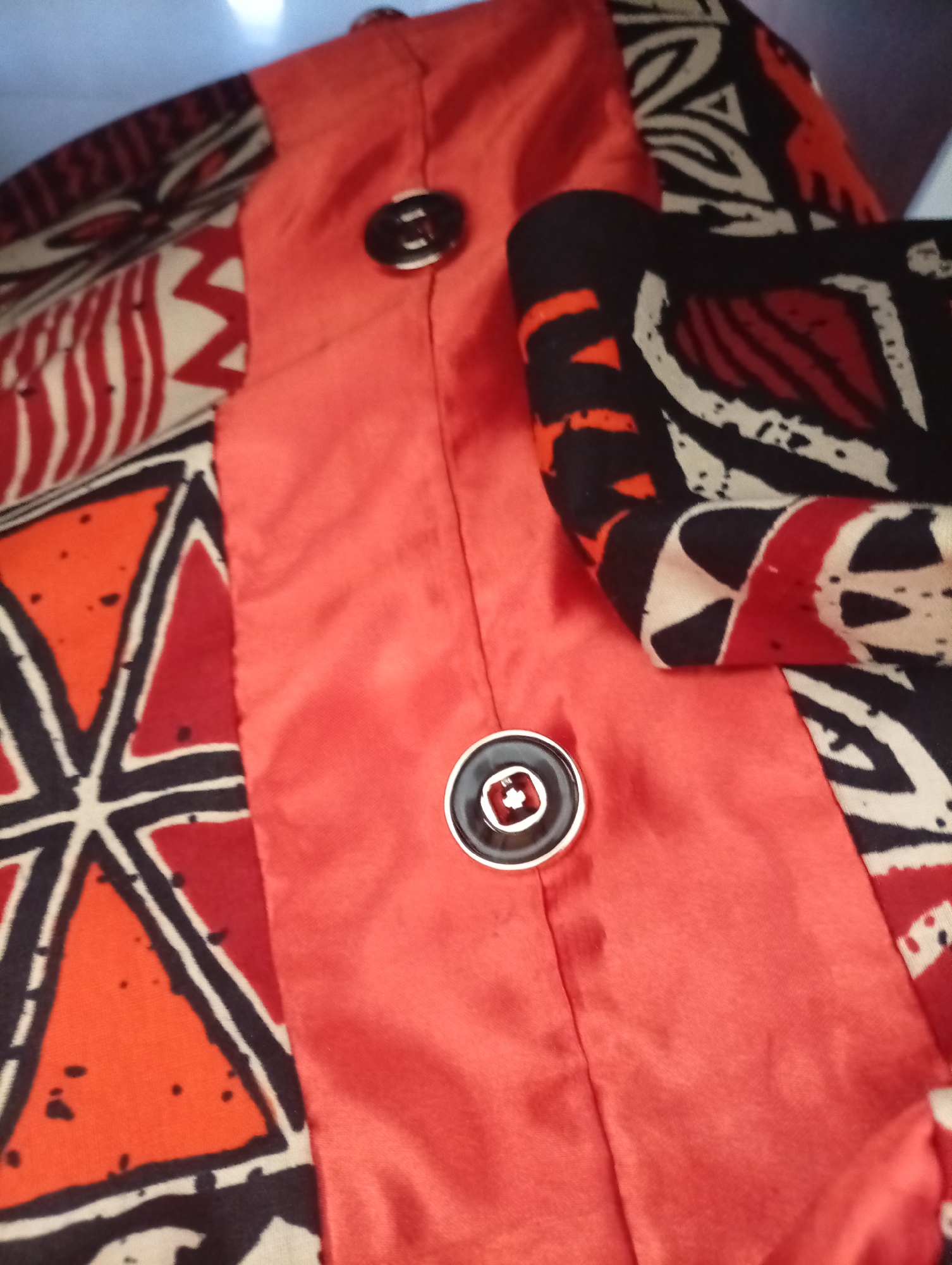 The image presents a close-up view of a vibrant garment, likely a jacket or top, featuring a combination of distinct fabrics and patterns. A prominent section of the clothing is made from a bright red or orange fabric with a smooth, satin-like sheen, suggesting a luxurious texture. A vertical seam runs down this solid-colored panel, adorned with two round buttons. Each button is black with a metallic silver or white outer ring; the lower button clearly displays a small, white, cross-like design in its center.
Adjacent to the solid red fabric are sections of textiles printed with bold, geometric, and abstract patterns. These patterns feature a striking palette of black, red, orange, and off-white or cream. Notable designs include sharp zig-zags, interconnected triangles, and stylized organic motifs, characteristic of traditional African textile prints. The patterned fabric appears to be a different texture, possibly cotton or a blend, providing a contrast to the smooth, solid red material.
The image is a static shot, focusing entirely on the details of the garment's construction and fabric. No people are visible, and no specific activity is taking place. The background is softly blurred, emphasizing the texture and design of the clothing. The style and prints on the fabric offer a strong cultural cue, consistent with the context of Sardauna, Nigeria, where such designs are common. There is no visible text in the image. The lighting appears even, highlighting the colors and textures without harsh shadows.