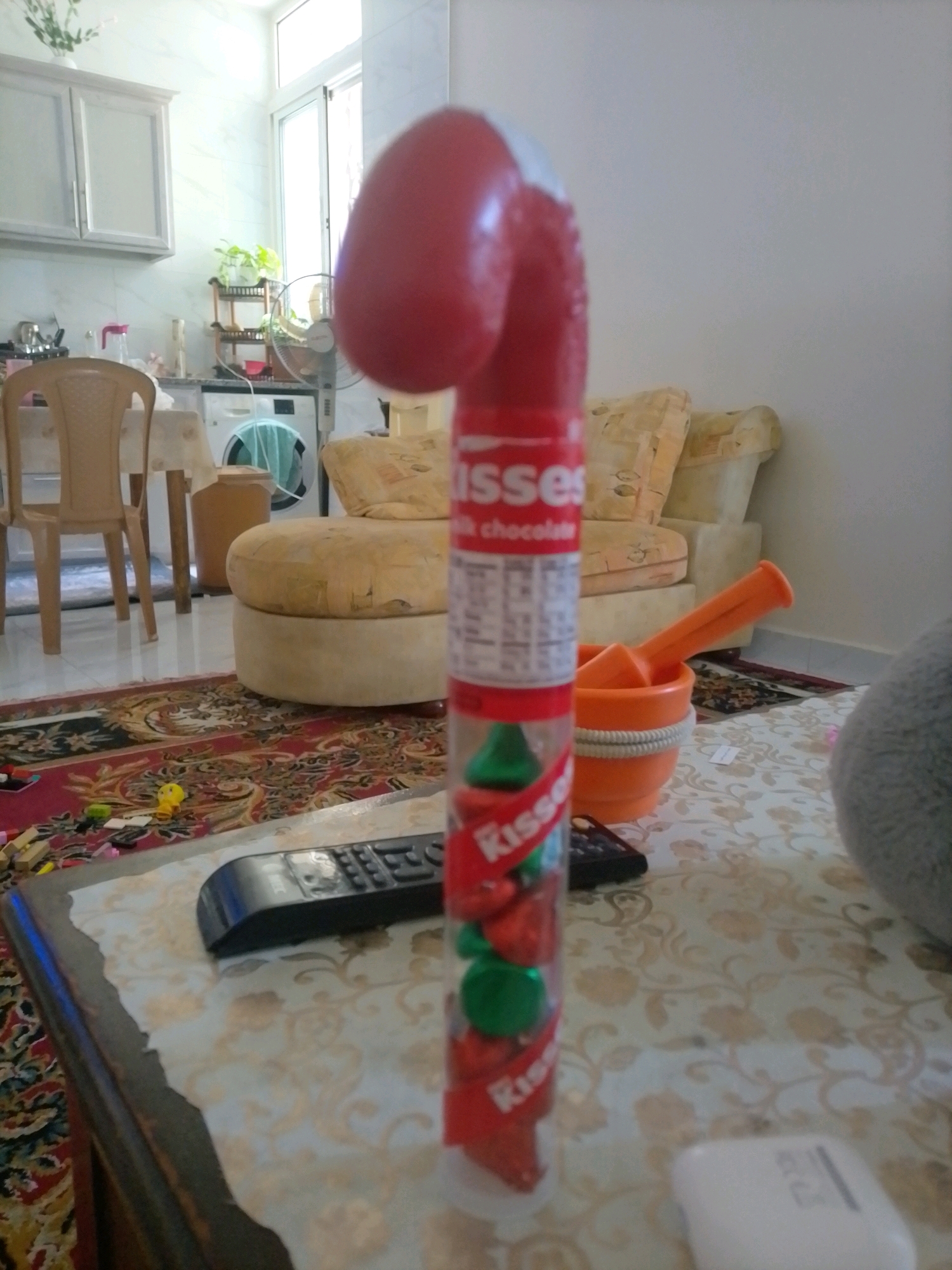 A candy cane-shaped dispenser for Hershey's Kisses, with a red curved top and a clear plastic tube filled with Kisses, stands on a patterned tablecloth. The Kisses inside are a mix of red and green, and the tube has "Kisses" printed on it. To the left of the candy dispenser, a black TV remote control rests on the tablecloth. In the background, a beige armchair and a sofa are visible, with a patterned rug on the floor. A washing machine and kitchen cabinets are also present in the background, suggesting a home environment. The lighting is bright, indicating daytime. The scene appears to be indoors, likely in a living room or dining area. There are no people visible in the image. The overall impression is a still life arrangement of candy and household items.