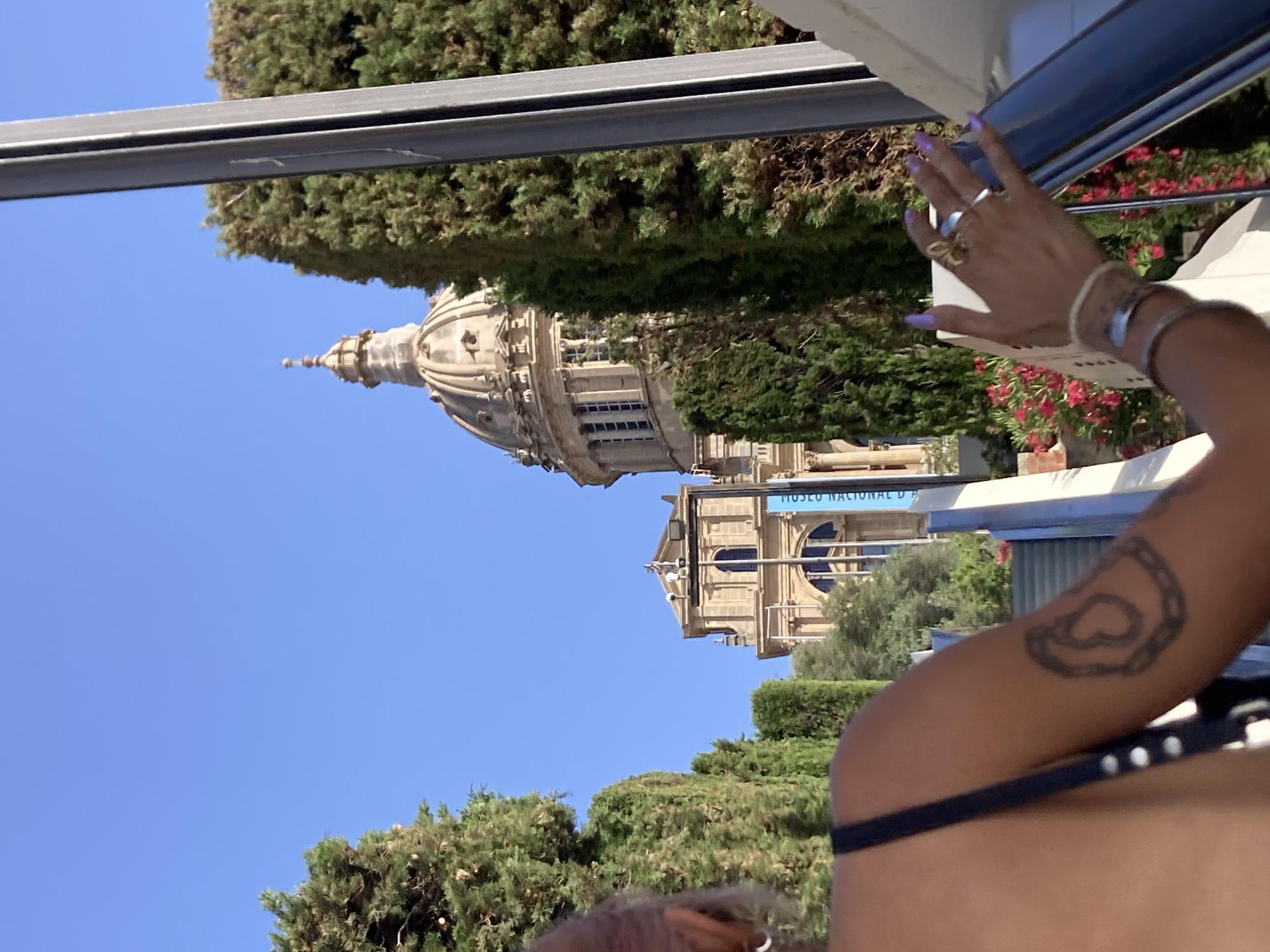 The image shows a view looking up towards the Palau Nacional in Barcelona, Spain. In the foreground, a person's shoulder and arm are visible, with a tattoo on their arm. The person is on an escalator, with their hand resting on the railing. The Palau Nacional is a large, ornate building with a dome, and it is surrounded by trees. The sky is clear and blue.