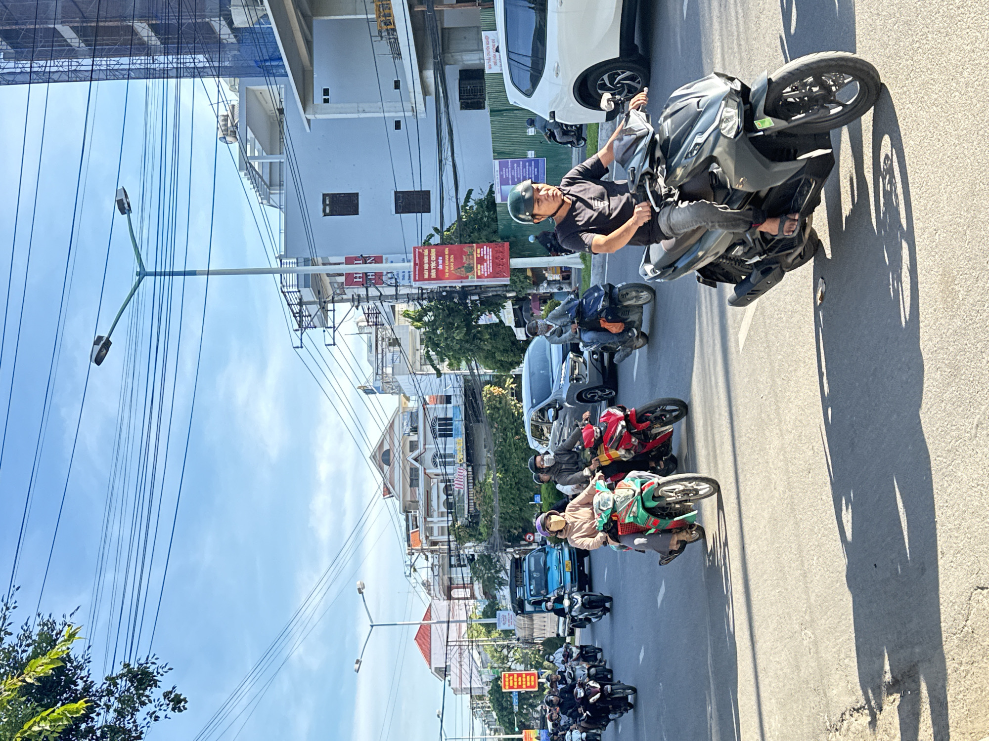 This is a daytime, eye-level, medium shot of a busy street scene in Nha Trang, Vietnam, featuring numerous motorcycles and a few cars. In the foreground, a man wearing a black t-shirt, gray jeans, and a dark helmet is riding a dark gray motorcycle. To his left, a woman in a light brown jacket, dark pants, and a patterned face mask is riding a green and black motorcycle, with a passenger behind her. To the right of the woman, a red motorcycle carries a driver. The background shows a two-lane road with traffic in both directions. Several other motorcycles, a blue truck, and a white SUV are visible. Buildings line the right side of the street, including a partially constructed building with blue netting. Power lines crisscross the sky, which is partly cloudy. Trees and streetlights are also present. The overall atmosphere suggests a typical urban street scene in Vietnam.