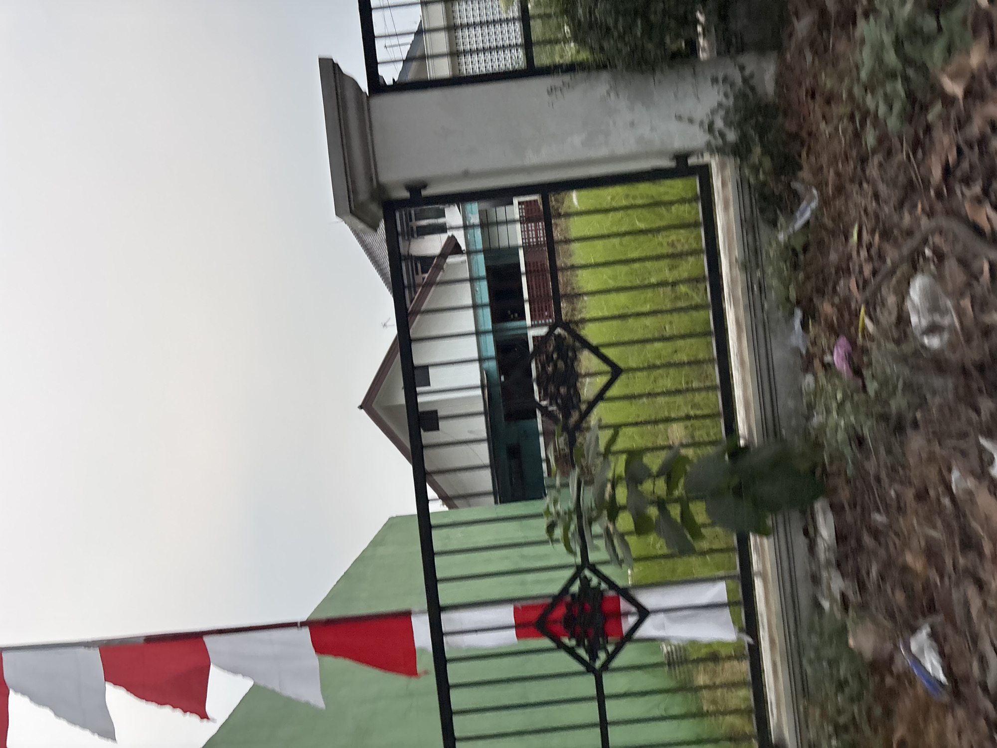 The image shows a fence in front of a building. A flagpole with a red and white flag is visible on the left side of the image. The building appears to be a house with a dark roof and white walls. There is grass in front of the building, and some trees and bushes. The sky is overcast. The scene is located in the city of Bekasi, Indonesia.