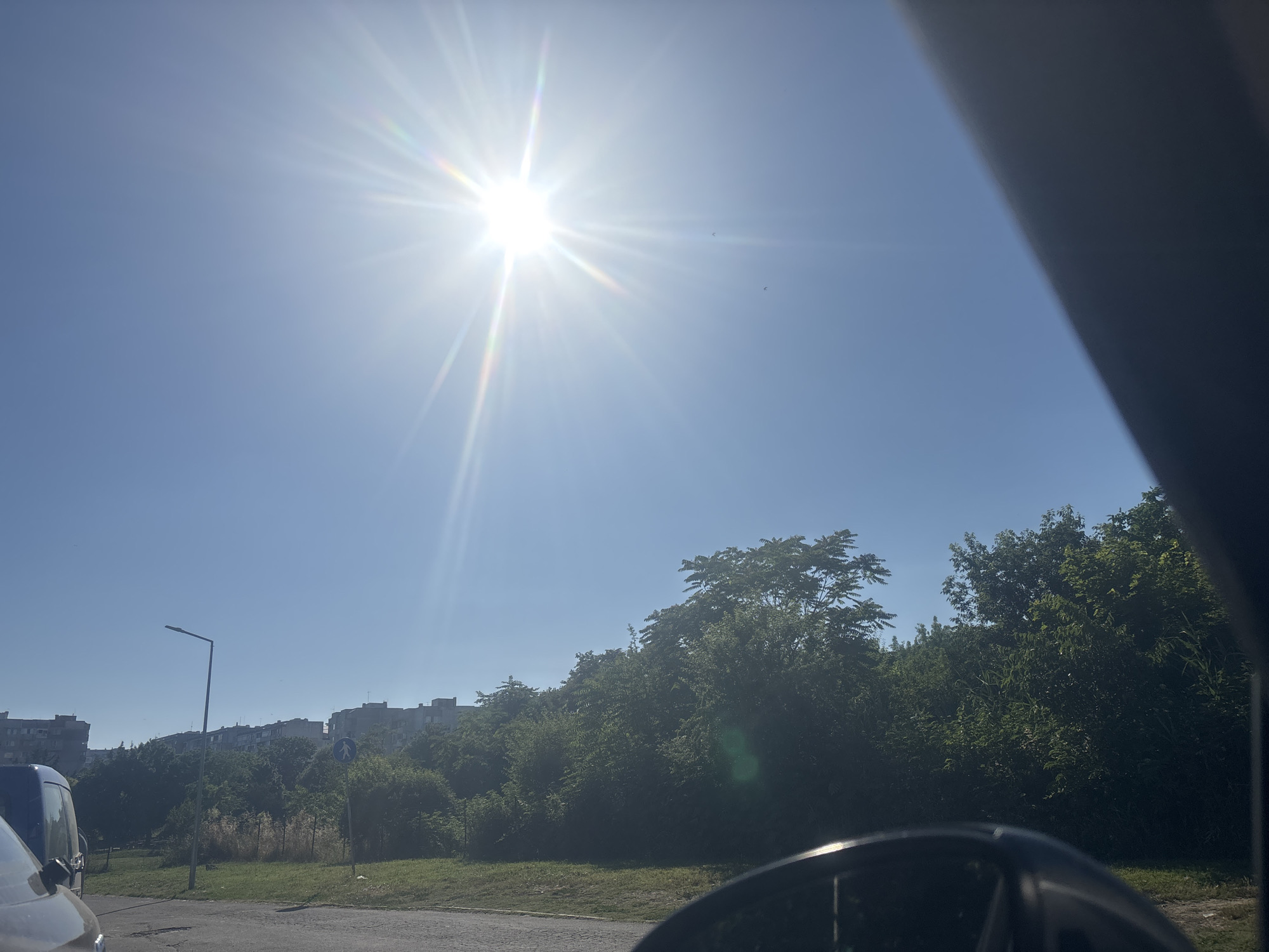 The image captures a bright, sunny day in Burgas, Bulgaria. The sun is high in the sky, casting strong rays and creating a lens flare effect. The scene appears to be a residential area, with apartment buildings visible in the background, nestled behind a dense line of green trees. A street lamp stands prominently on the left side of the frame, and a pedestrian crossing sign is visible amidst the foliage. The foreground shows a paved road and a grassy verge. Parts of vehicles are visible in the lower section of the image, suggesting the photo was taken from inside or near a car. The clear blue sky and intense sunlight indicate it is midday on a warm day. No specific activity or event is depicted, but the setting suggests a typical urban or suburban environment in Bulgaria.
