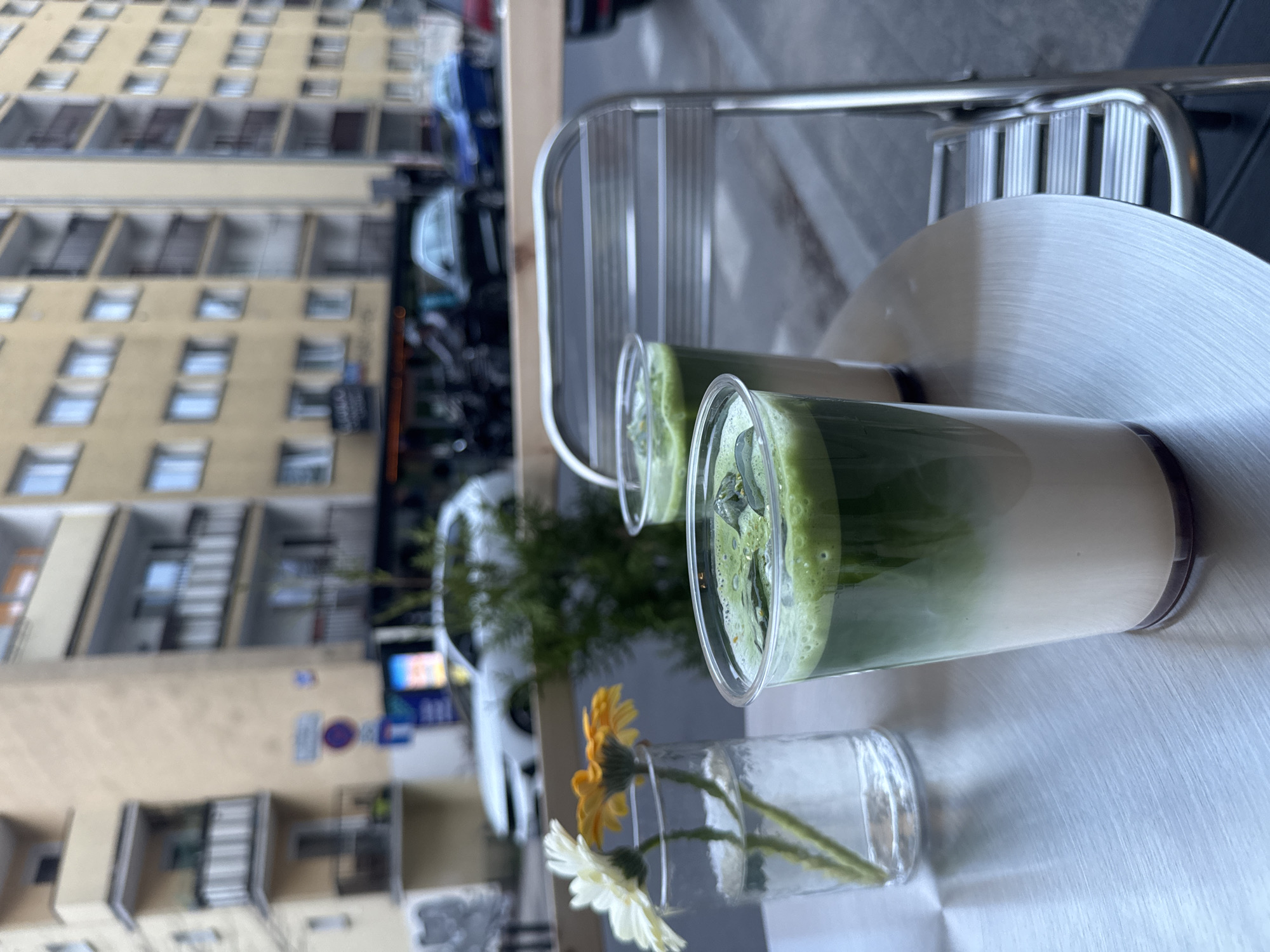 Two transparent plastic cups, containing layered beverages of white and green liquid, sit on a light-colored metallic table. A small, clear glass vase with a few yellow and white daisies is also on the table. Behind the cups and vase are two metal chairs with slatted backs. In the blurred background, a multi-story building with numerous windows is visible, as well as parked cars and street signs. Based on the architectural style and street signs, the location is likely Warsaw, Poland.