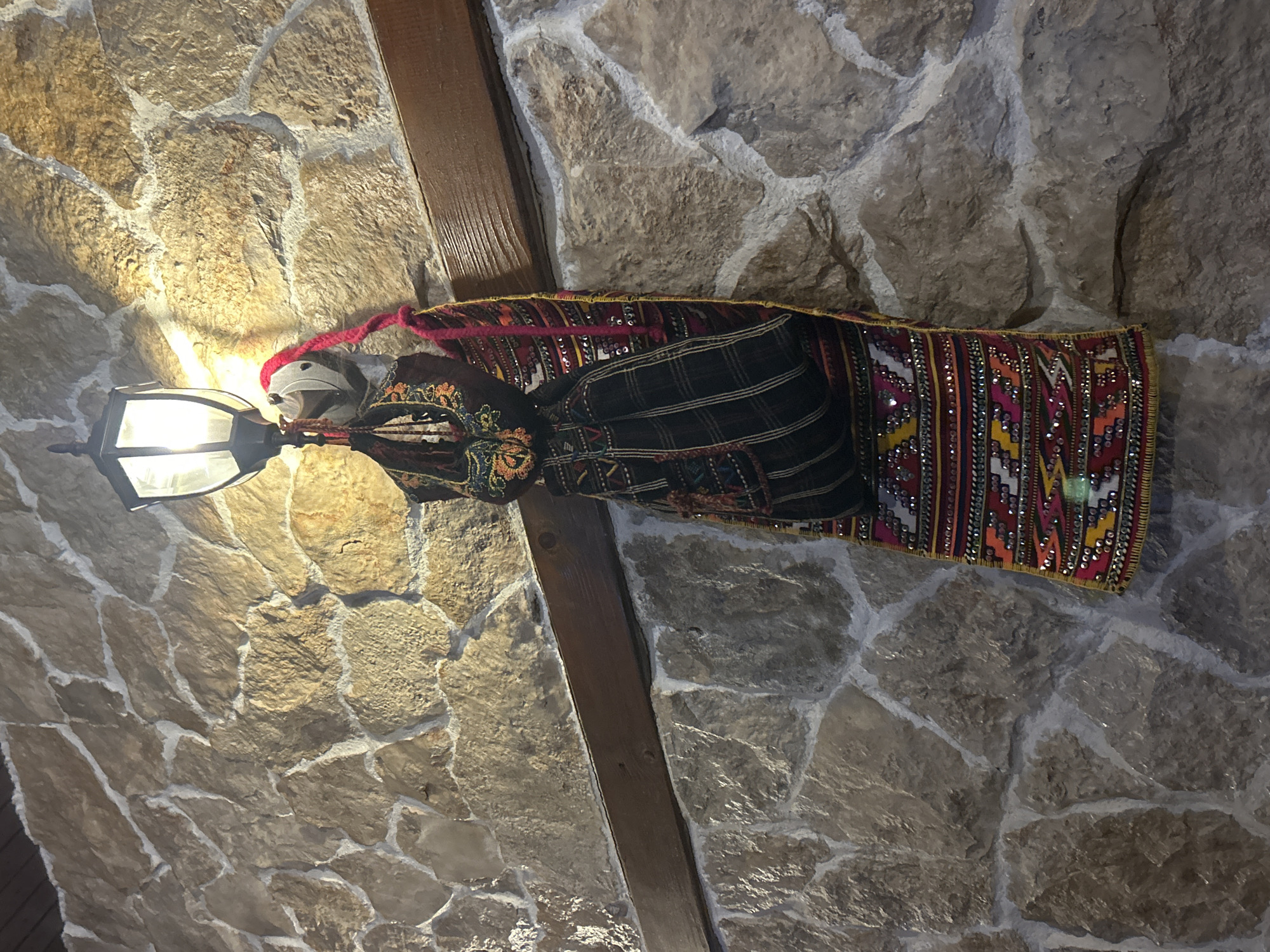 The image shows an interior wall constructed of irregularly shaped stones of light beige and gray. A dark wooden beam runs diagonally across the wall. Suspended from a light fixture attached to the wall is a decorative, possibly hand-woven, textile object. The textile exhibits a complex pattern with various colors, including red, black, and gold, and is adorned with sequins. The light fixture has a classic lantern design and is lit, casting light on the surrounding stone wall. The setting might be a building interior in Mirovyane, Bulgaria, considering the mention of the city.