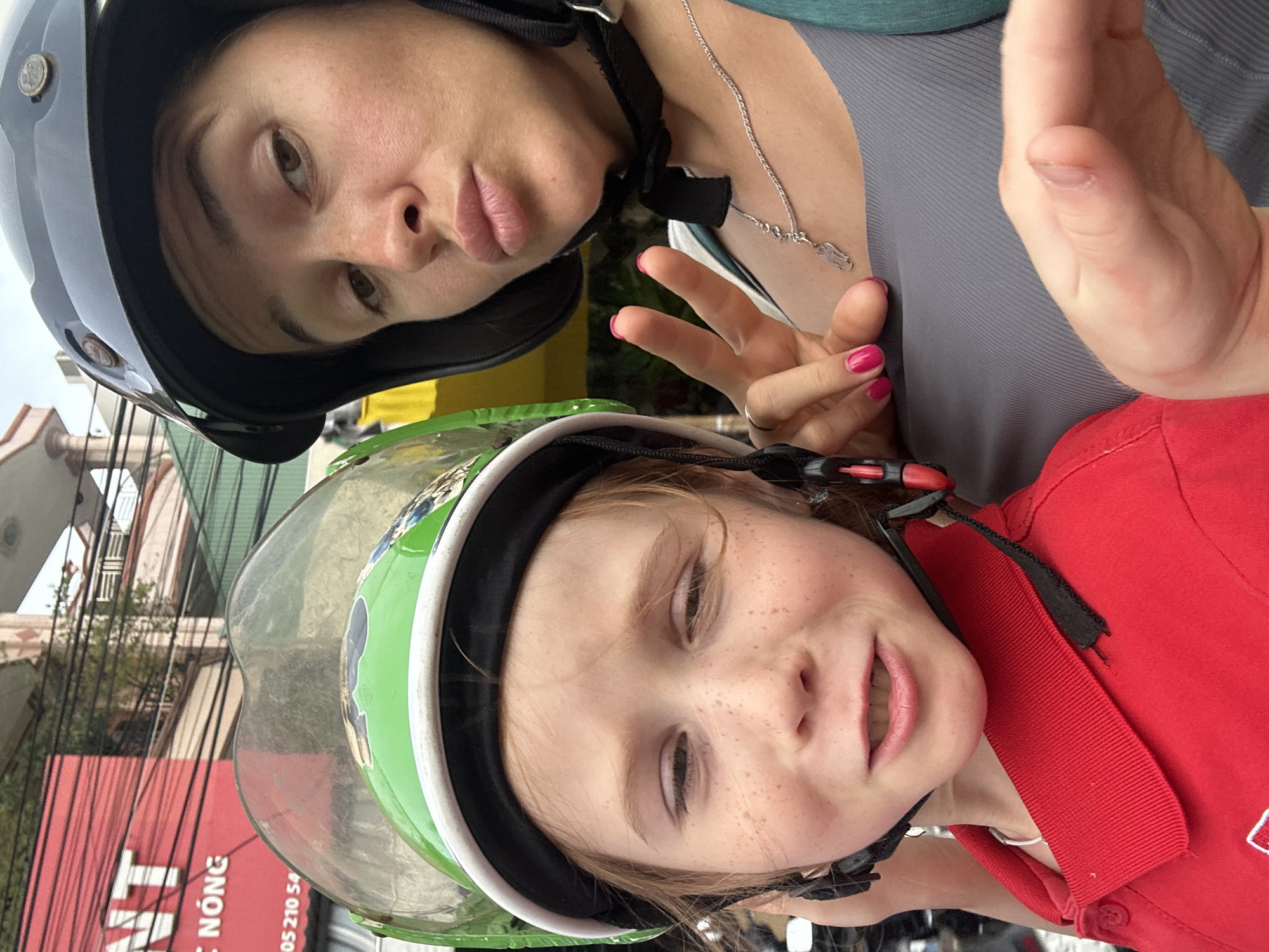 Two people, a woman and a child, are posing for a selfie on a motorbike in Bac Nha Trang Ward, Vietnam. The woman, wearing a black helmet and a grey tank top, puckers her lips and makes a peace sign with her right hand. She is wearing a silver necklace with a small charm. The child, wearing a green helmet and a red polo shirt, looks at the camera with a slightly squinted expression. Both are wearing helmets for safety. In the background, a red sign with white lettering is visible, along with buildings and trees, indicating an urban setting. The lighting suggests it is daytime.