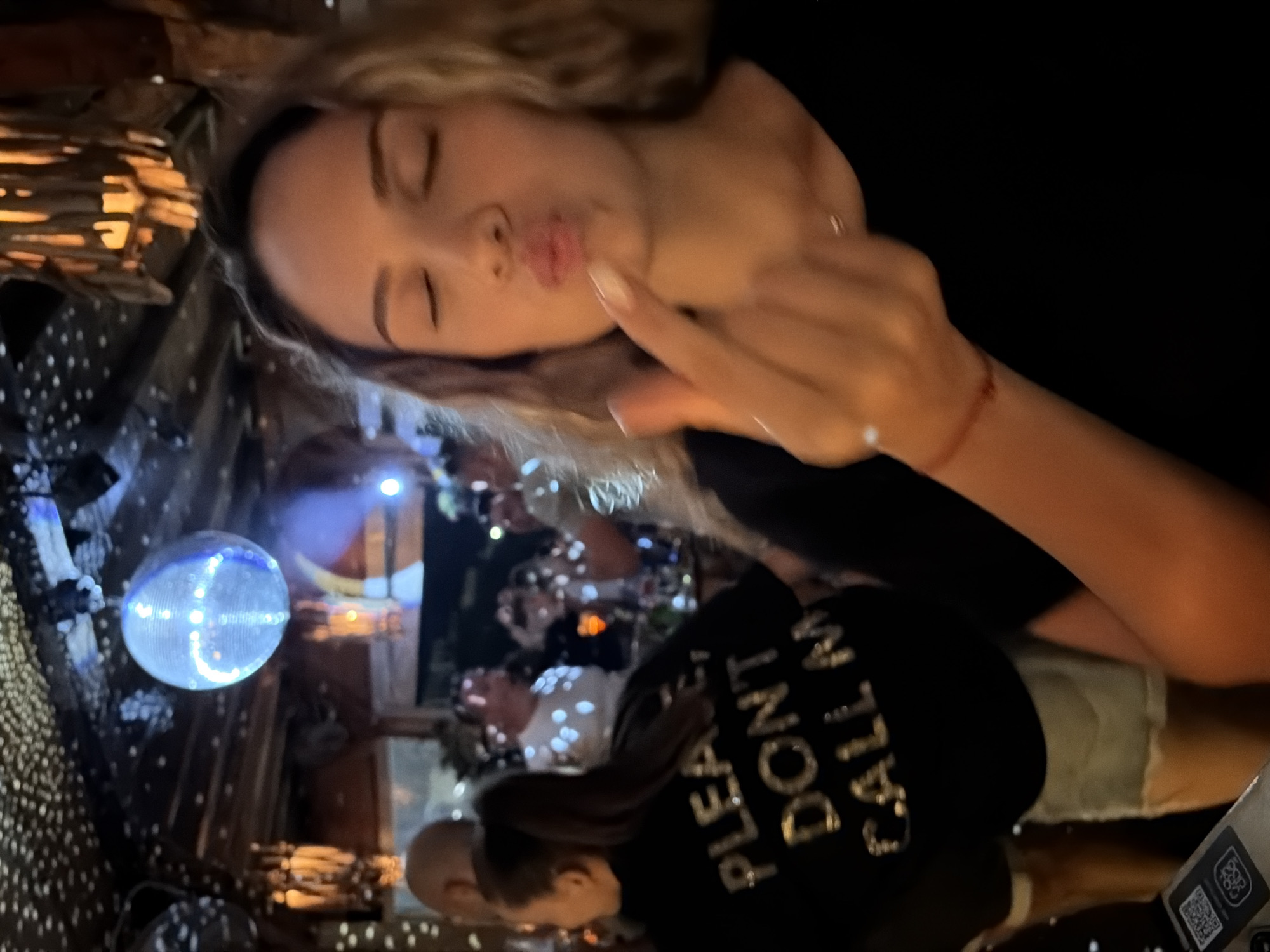 A young woman with blonde, curly hair is pictured from the chest up, closing her eyes and puckering her lips while extending her right index finger towards the camera. She is wearing a black t-shirt with a silver-sequined text on the back that reads "PLEASE, DON'T CALL M". She has a delicate necklace around her neck and a thin red bracelet on her left wrist. The background suggests a lively indoor environment, possibly a bar or club, with a disco ball hanging from the ceiling and warm lighting emanating from decorative lamps. Other people are visible in the background, some appearing to be dancing or socializing. The setting is identified as Lozenets, Bulgaria.