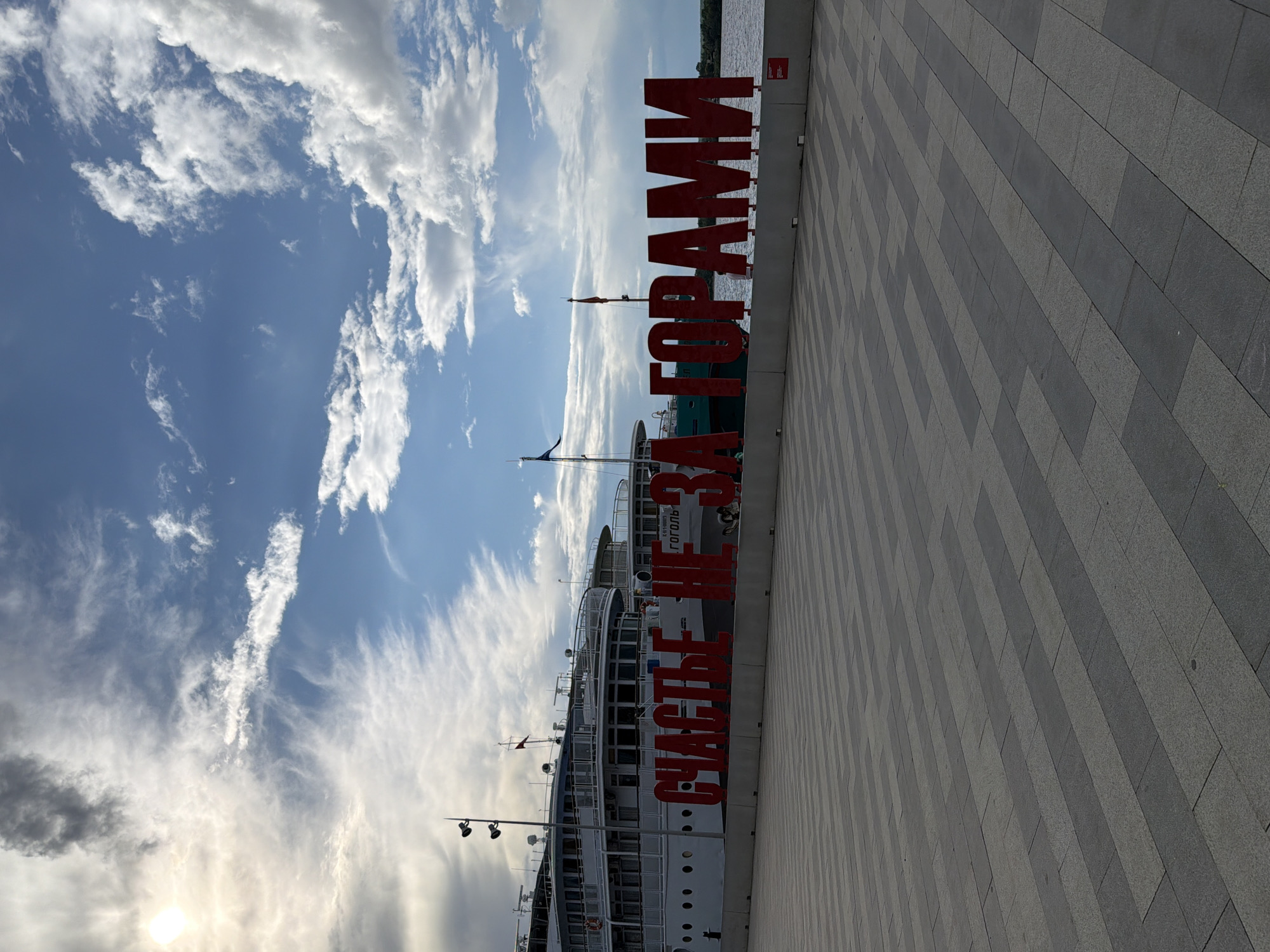 This is a photograph taken in Perm, Russia, featuring a large red sign with Cyrillic text that reads "СЧАСТЛИВЫМ ГОРОДАМ" (To Happy Cities). The sign is placed along a waterfront promenade paved with grey and beige tiles. In the background, a large white ship with multiple decks is docked. The sky is a vibrant blue with scattered, wispy white clouds, suggesting it is daytime with fair weather. The sun is visible in the lower left corner, partially obscured by clouds, indicating it might be late afternoon. The scene appears to be a public space, likely a tourist or recreational area, given the promenade and the presence of a ship. The overall mood of the image is bright and optimistic, reinforced by the "happy cities" message.