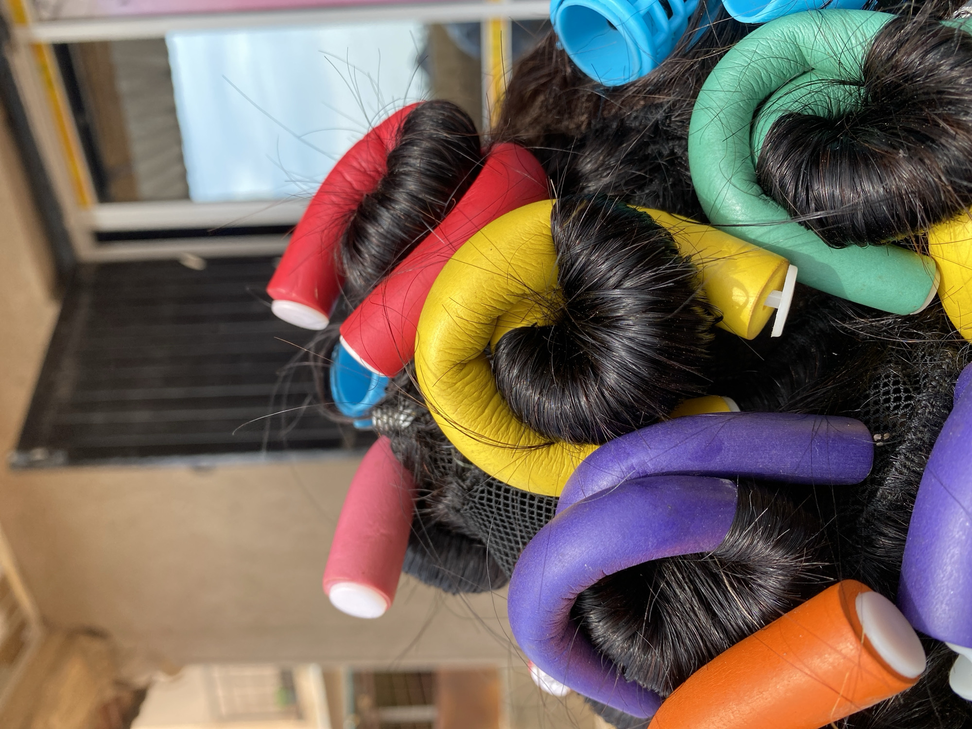 This is a close-up, eye-level shot of a person's hair, covered in colorful foam rollers, in what appears to be an outdoor setting in Bar Jirgi Summa, Nigeria. The hair is dark and appears to be styled in large buns. The rollers are of various bright colors: red, pink, orange, purple, yellow, and green. Some blue plastic rollers are also visible, one on the right and another partially visible on the left. The rollers are wrapped around sections of hair, in the process of creating curls. The background is blurred but suggests an outdoor environment with a building and possibly a window or door with vertical bars. The lighting indicates it is daytime, with sunlight casting subtle shadows. The overall impression is a snapshot of a hair styling process taking place outdoors.