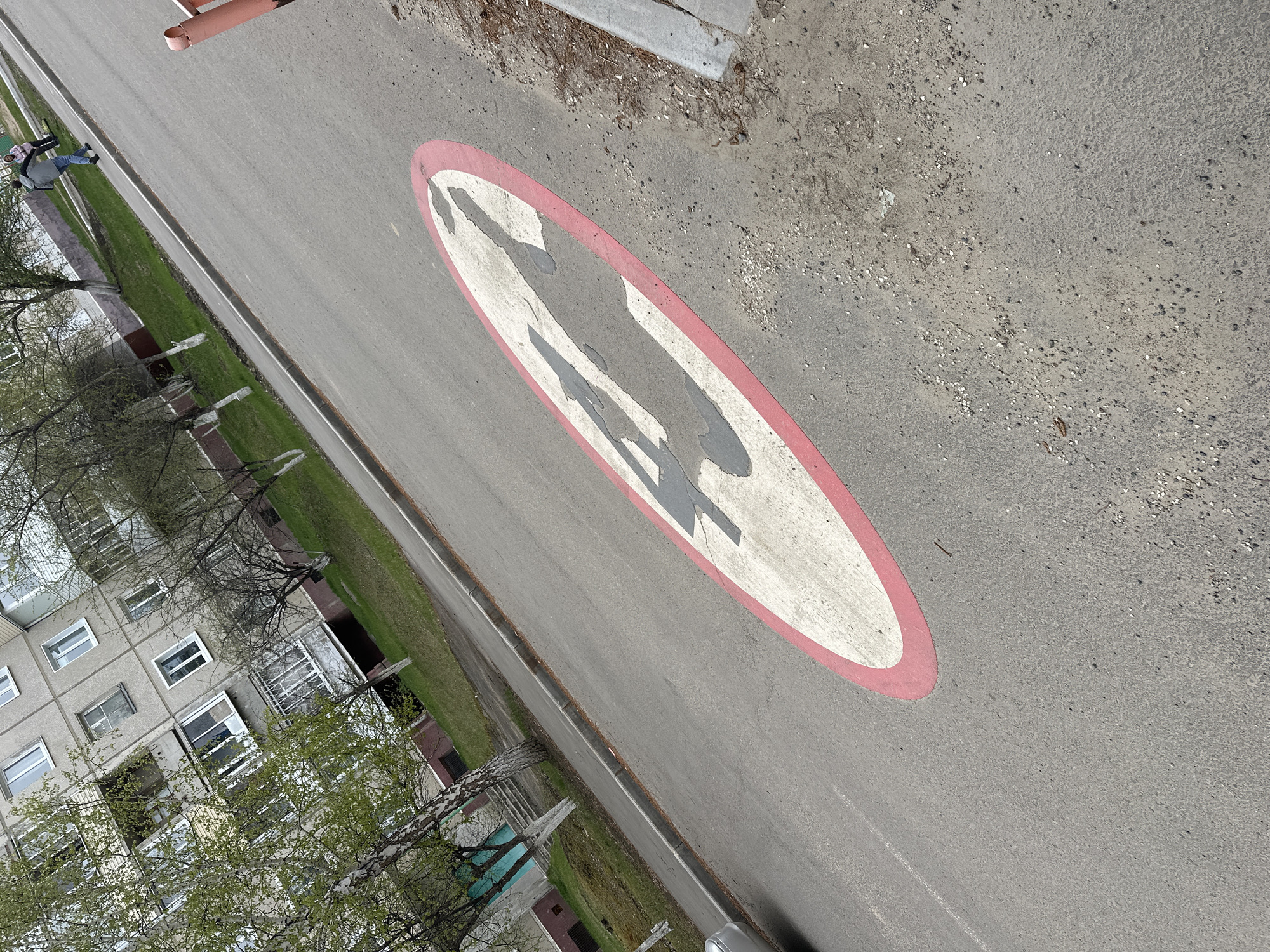 The image shows a street in Sayansk, Russia. The street has a faded road marking depicting pedestrians crossing. A multi-story residential building is visible on the left, partially obscured by trees. Two people are seen walking along the side of the road. The street surface is gray asphalt with some dirt and debris.