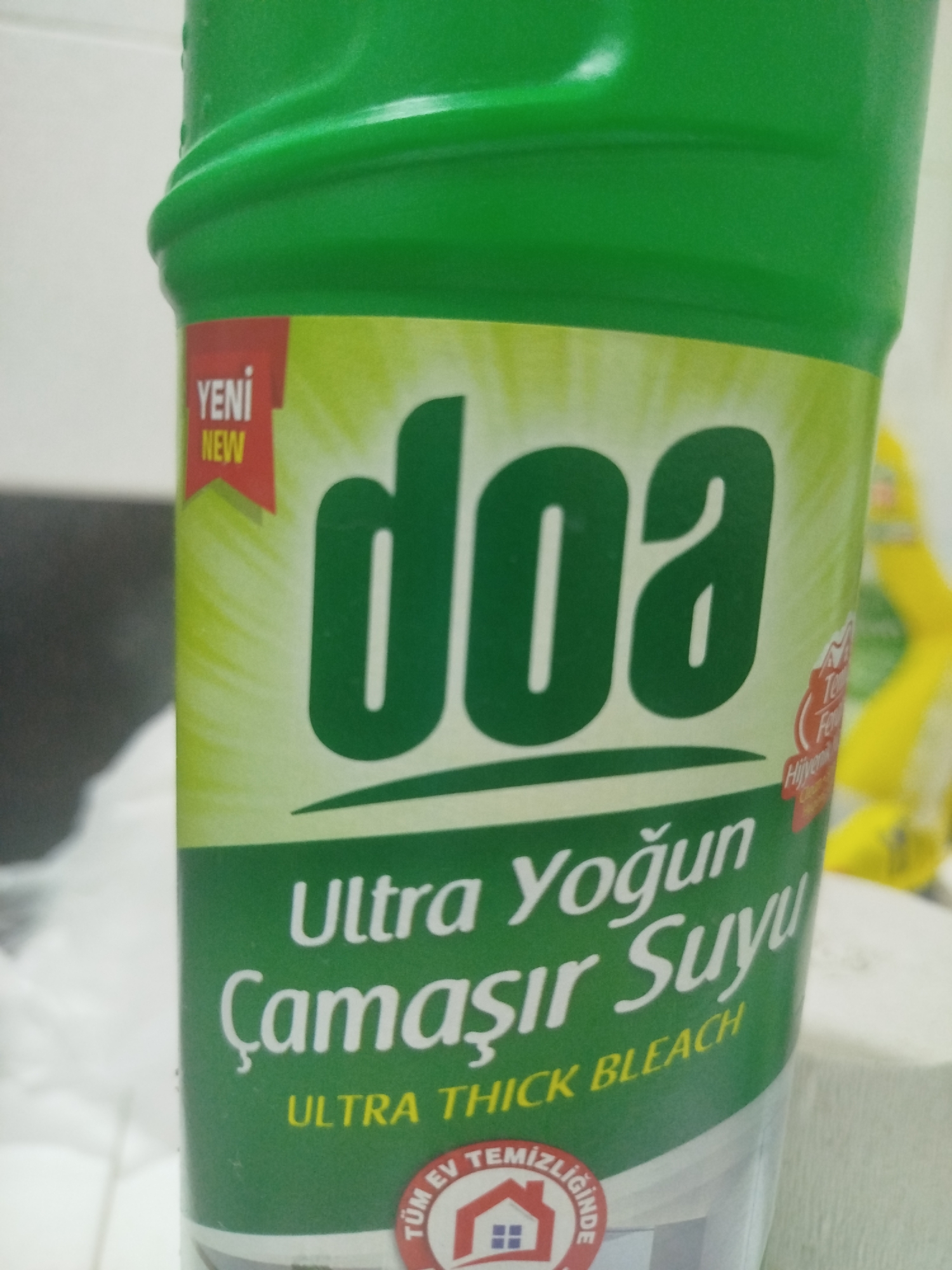 The image shows a close-up of a green plastic bottle of cleaning product. The label on the bottle is predominantly green and yellow, with white text. Prominently displayed is the brand name "doa" in large green letters. Below this, the text reads "Ultra Yoğun Çamaşır Suyu" which translates to "Ultra Concentrated Laundry Water" or "Ultra Concentrated Bleach". Further down, in smaller yellow letters, it says "ULTRA THICK BLEACH". A red tag with "YENİ NEW" is visible on the left side of the bottle. The background is blurred, but appears to be an indoor setting with a white surface and a hint of yellow in the background. There are no people or animals visible in the image, nor is there any indication of the city of Batumi, Georgia. The image is a product shot focused solely on the cleaning product.