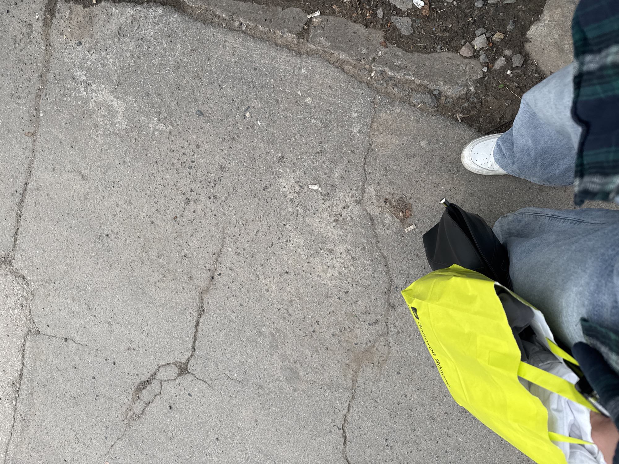 A first-person perspective shot shows a person standing on a cracked concrete sidewalk, likely in a city. The person is wearing light blue jeans and a white sneaker. To the left, a bright yellow shopping bag is visible, with a black bag next to it. The ground is littered with small debris, including what appears to be cigarette butts and small stones. The overall environment suggests an outdoor urban setting. The yellow bag has text in Russian, which reads "золотое яблоко", meaning "Golden Apple", a popular beauty retailer in Russia, including in the city of Perm.