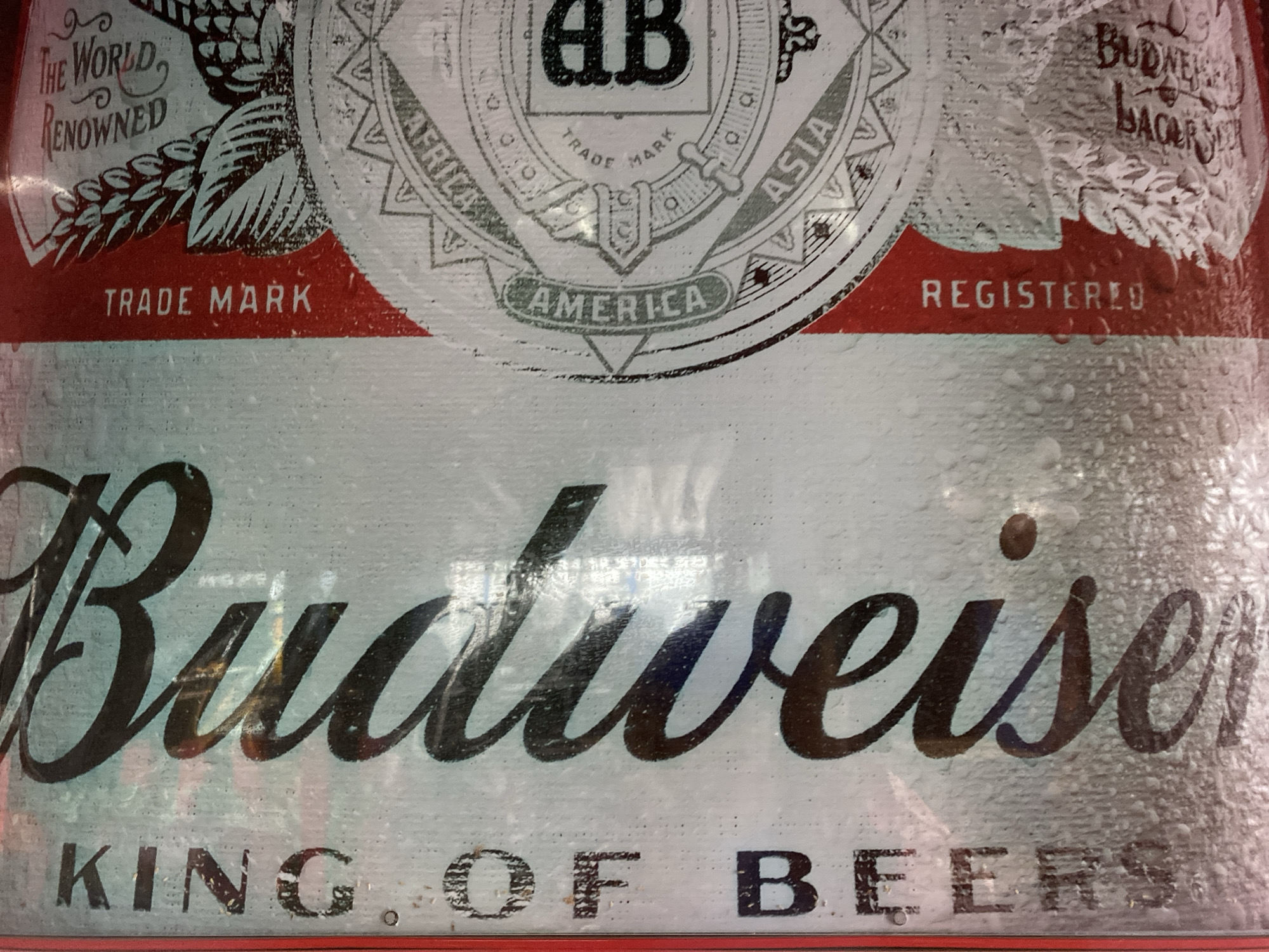 The image is a close-up shot of a Budweiser beer can, likely taken in Howayi, Nigeria. The can features the distinctive Budweiser logo, with the script "Budweiser" prominently displayed in black. Below it, the slogan "KING OF BEERS" is also visible. The can appears to be cold and has water droplets on its surface, suggesting it has been refrigerated. The background is blurred, but hints of red and white, the brand's signature colors, are present in the upper portion of the can, which displays additional branding elements such as "TRADE MARK," "REGISTERED," and potentially part of a shield or crest with text like "AFRICA," "ASIA," and "AMERICA." The overall impression is that of a product shot, focusing on the branding and visual appeal of the beer can. There are no people or any specific environmental details of Howayi, Nigeria, discernible from the image itself.