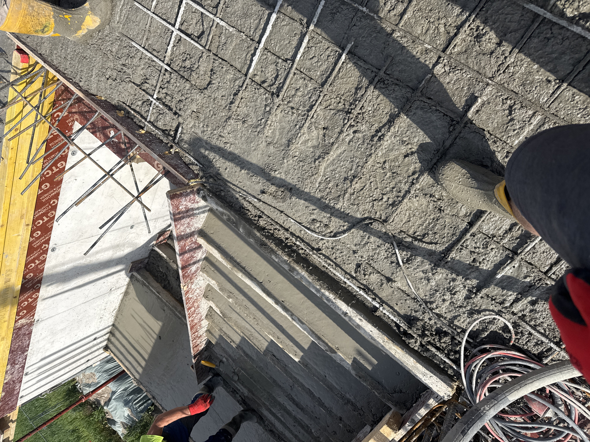 The image depicts a construction site, likely in the process of pouring concrete. A partially completed concrete structure is visible, reinforced with a steel rebar grid. Fresh, wet concrete covers a large area, with footprints and shadows suggesting recent activity.
Formwork, constructed from wooden planks and yellow support beams, defines the edges of the structure. A worker in a blue uniform and red gloves is visible, seemingly involved in smoothing or finishing the concrete. Electrical cables are coiled nearby, suggesting the use of power tools or equipment.
The image doesn't provide explicit information to confirm the location as Plovdiv, Bulgaria, and no landmarks or signs are visible to confirm it.