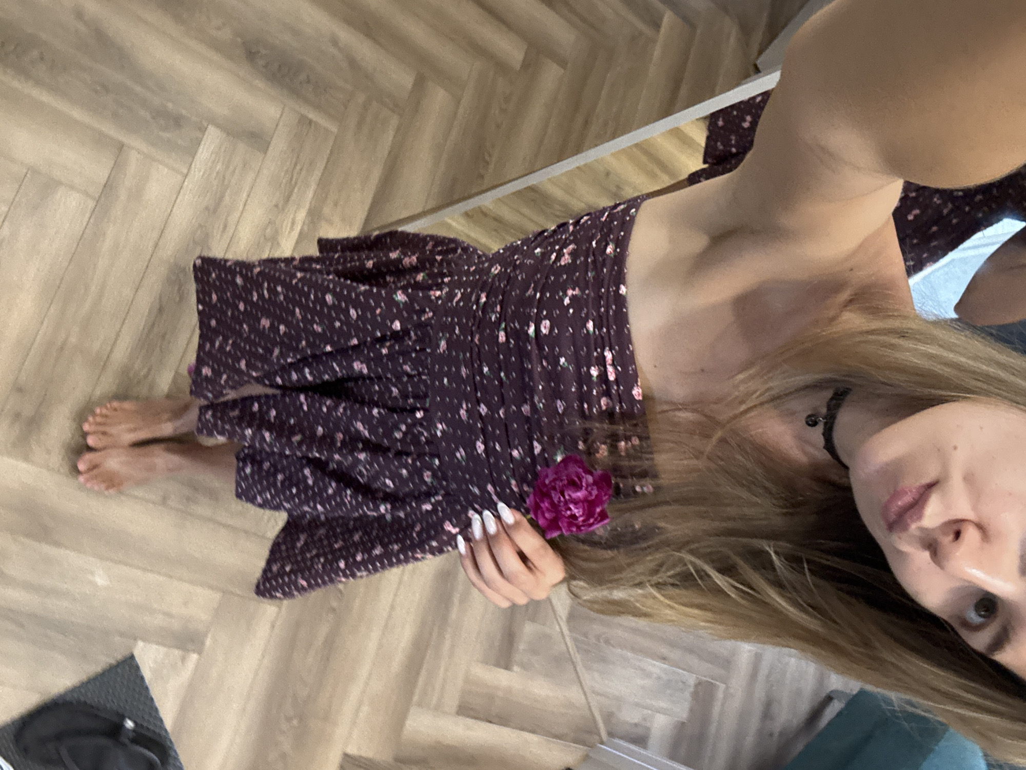 A young woman with blonde hair is taking a selfie while wearing a floral print dress and a black choker necklace. She is holding a pink flower in her right hand. The woman is standing in what appears to be an indoor setting, possibly a room with wooden flooring and a mirror reflecting part of her. Her feet are bare. In the background, there is a hint of a teal-colored furniture item. The overall setting is ambiguous, with no clear indicators of a specific city or outdoor environment.
