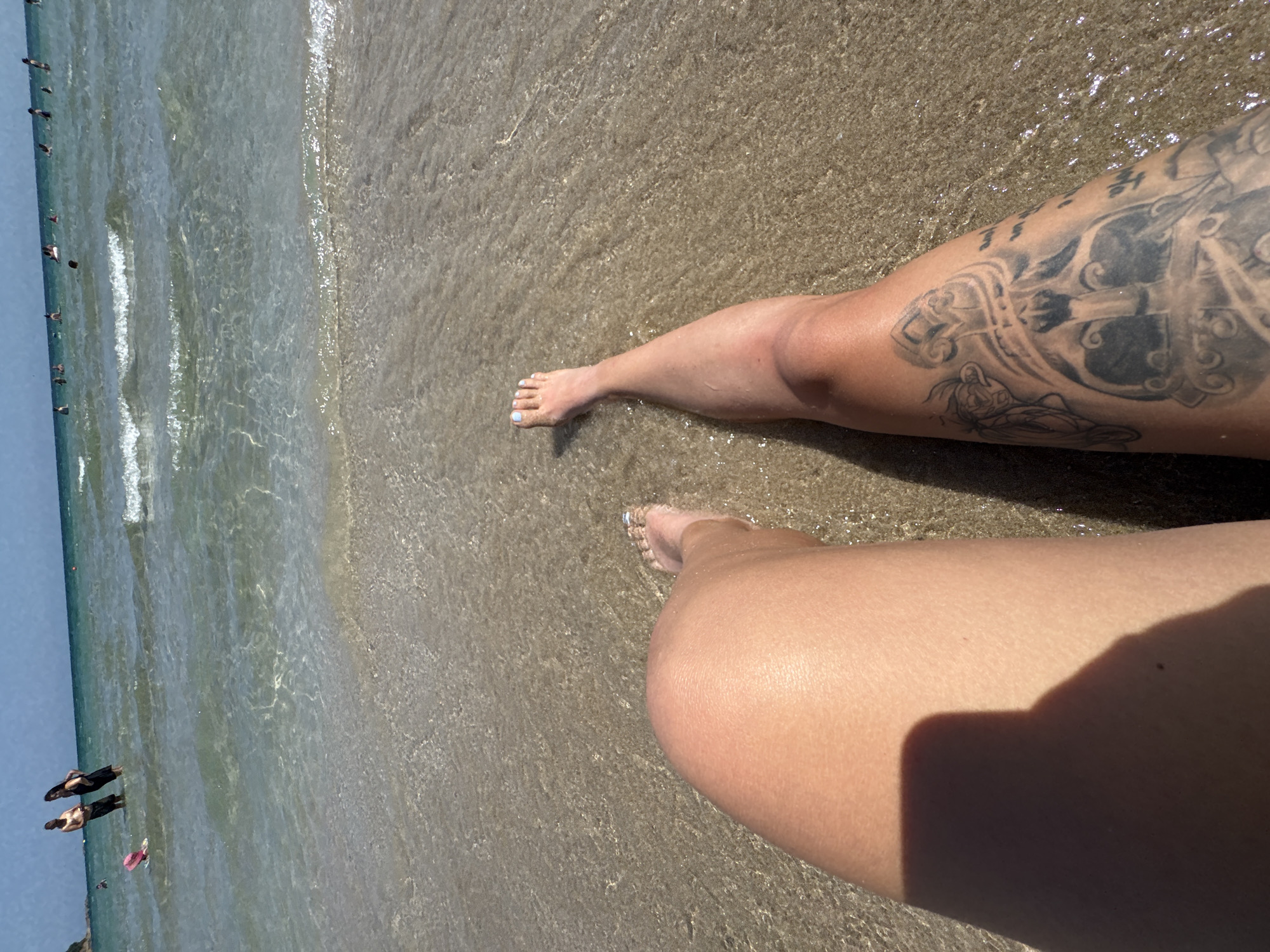 The image shows a first-person perspective of someone sitting in shallow water at a beach. The foreground is dominated by the person's legs, one with a large tattoo of what appears to be a helmet with intricate designs and some script. The other leg is bare. Both feet are visible with painted toenails. The water is clear and shallow, with sand visible beneath the surface. In the background, there are several people in the water and on the shoreline. Some are wading, while others appear to be swimming or standing. The sky is clear and blue, and the overall scene suggests a sunny day at the beach. The location is identified as Primorsko, Bulgaria.