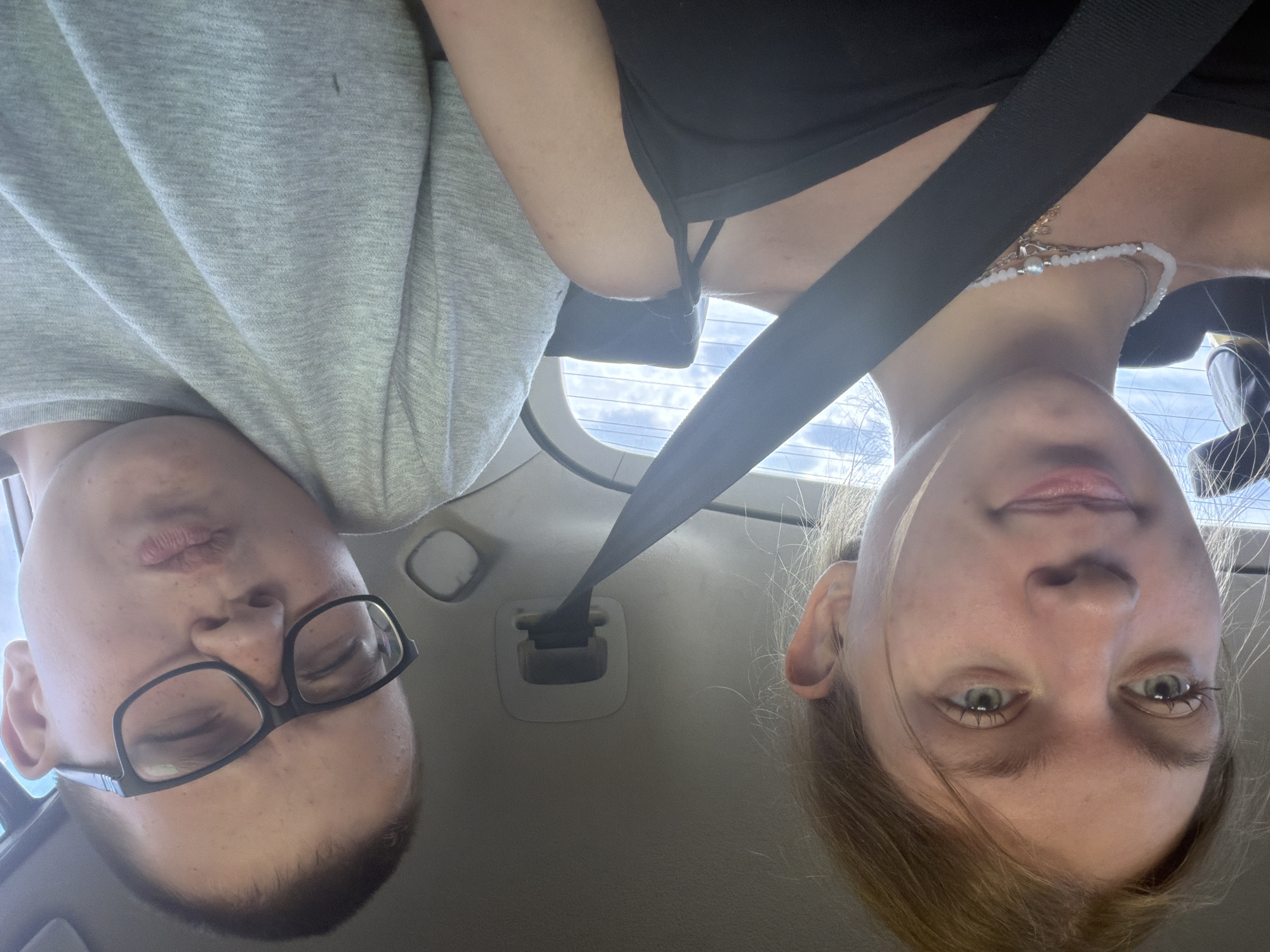 Two individuals are in the back seat of a vehicle, with a seatbelt extending between them. The person on the left, appearing to be a young female with blonde hair, looks towards the camera. She is wearing a black tank top and a beaded necklace. The person on the right, appearing to be a young male wearing glasses and a grey t-shirt, is looking towards the camera with a pouting expression. The background shows the interior of the car, including a seatbelt buckle mechanism and the car's rear window, which displays a cloudy sky. The image was captured in Ostrovka, Russia.