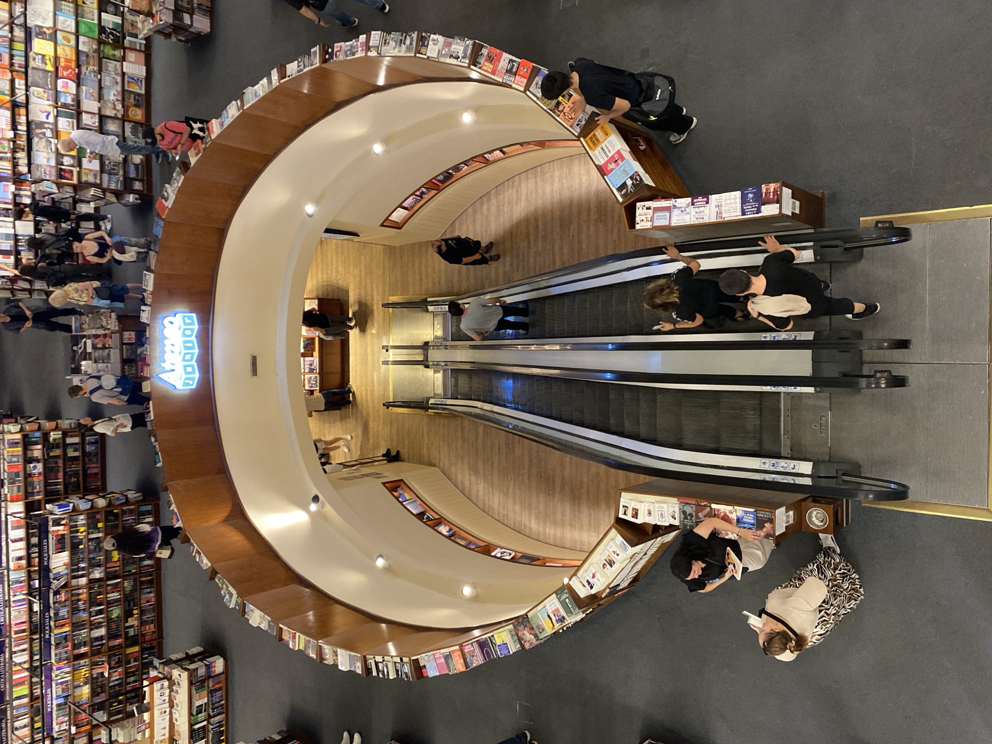 This overhead image depicts the interior of El Ateneo Grand Splendid bookstore, located in Buenos Aires, Argentina. The scene features a multi-level retail space with a central, semi-circular void revealing two parallel escalators descending to a lower level characterized by wooden flooring. This void is bordered by a curved, light brown wooden balustrade, which serves as a display surface for numerous books. Above the escalator entrance, a blue neon sign displays the text "Ateneo Junior."
The primary visible floor, appearing in the foreground and surrounding the central void, is carpeted in dark grey. It is populated by numerous tall, dark wooden bookshelves, extensively stocked with books and arranged in aisles. Additional bookshelves line the upper perimeter walls visible in the frame.
Approximately 25 individuals are distributed across the visible areas. Activities observed include browsing books on shelves and display units. Several individuals are utilizing the escalators: two are descending on the right escalator, while two others are ascending on the left. On the main floor, two women are interacting with books at a display stand in the lower-left quadrant. A man in a black shirt is holding a book near a display stand in the lower-right. Most individuals are dressed in casual attire. Recessed lighting fixtures illuminate the interior from the ceiling of the upper level.