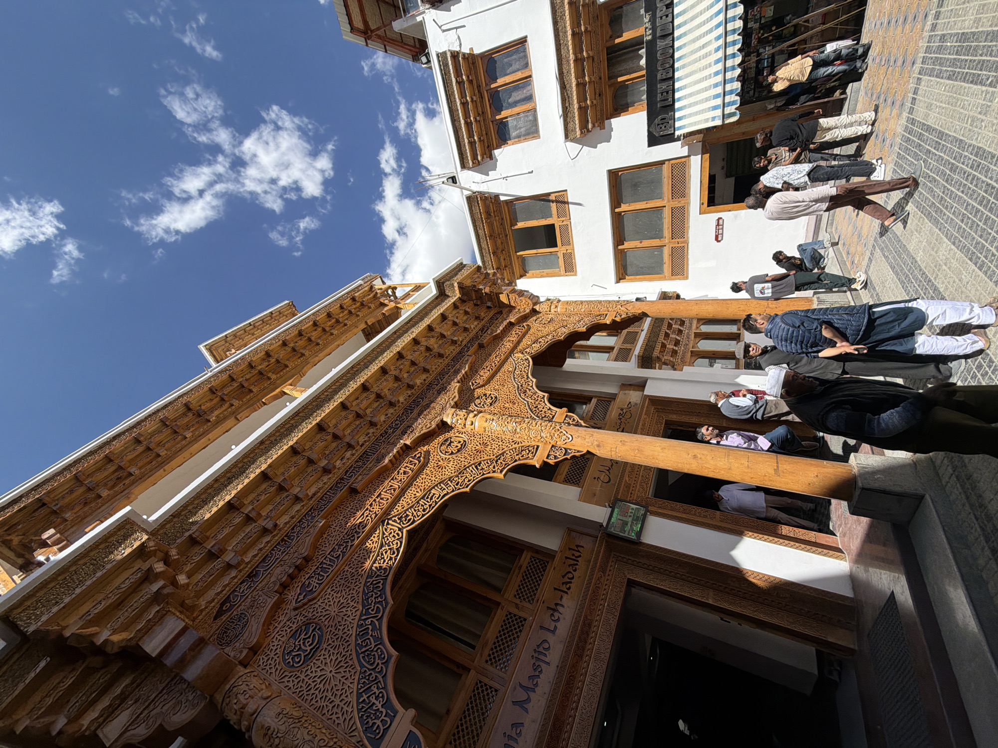 In Leh, India, the exterior of the Jama Masjid Leh-Ladakh is captured from a low angle, emphasizing its ornate wooden architecture. Sunlight illuminates the intricate carvings on the wooden facade, which features multiple arches and windows. To the right of the mosque, a white building with similar wooden window frames is visible, housing a store labeled "LEH BOOK DEPOT" with a striped awning. Several people are present in the scene, some entering or exiting the mosque, while others walk along the cobblestone path or gather outside the bookshop. The sky above is blue with scattered white clouds, indicating a clear day. The overall impression is a bustling street scene in front of a significant cultural landmark.