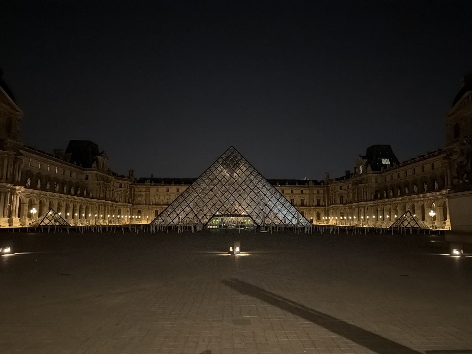 At night, the Louvre Museum and its iconic glass pyramid stand illuminated against a dark sky in Paris, France. The pyramid is centered and prominent, surrounded by the museum buildings on either side, which are lit by numerous lamps. A vast paved courtyard extends in front of the pyramid, with small, ground-level lights marking the space. The lighting creates a warm, golden hue on the building exteriors and the courtyard, contrasting with the darkness above. There are no people visible in the image.