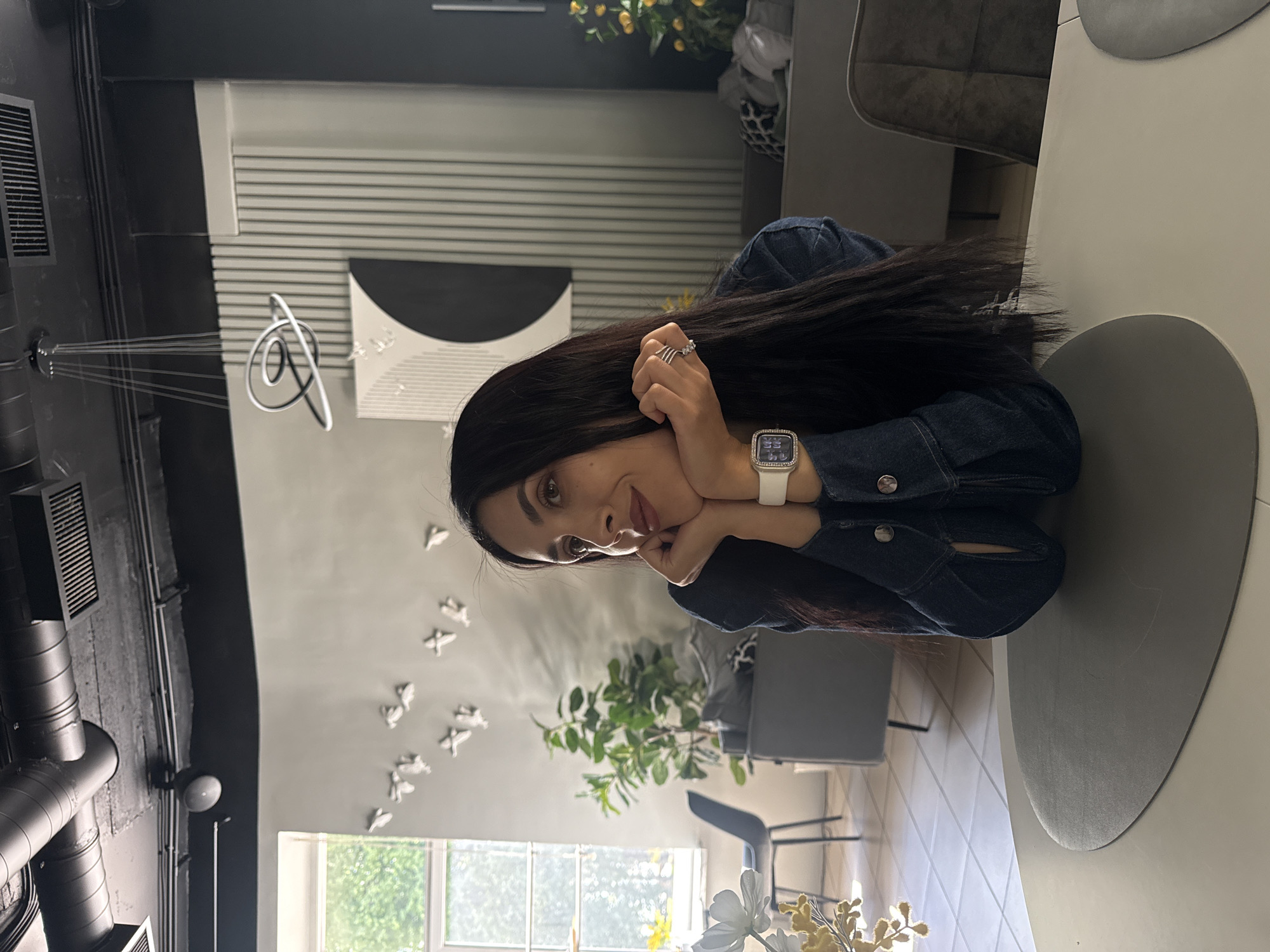 A young woman with long, dark hair is seated at a table, resting her chin on her hands. She wears a dark denim jacket and a white smartwatch. Her gaze is directed towards the camera, and she has a gentle smile on her face. The background suggests an indoor setting, possibly a cafe or restaurant, with modern decor. Abstract art pieces and a hanging light fixture are visible on the wall behind her. A window with a view of greenery is to her left. The lighting indicates it is daytime.