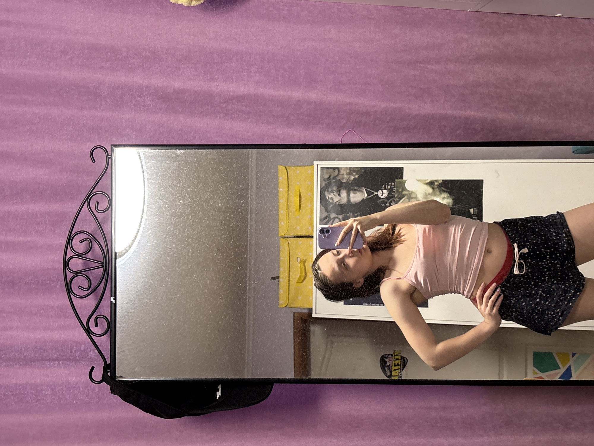 A young woman takes a selfie in a mirror. The mirror has a decorative wrought iron stand. The background wall is purple. A framed picture and a yellow object are visible in the background. The woman is wearing a pink tank top and dark shorts. The location cannot be determined to be Прикамский, Russia.