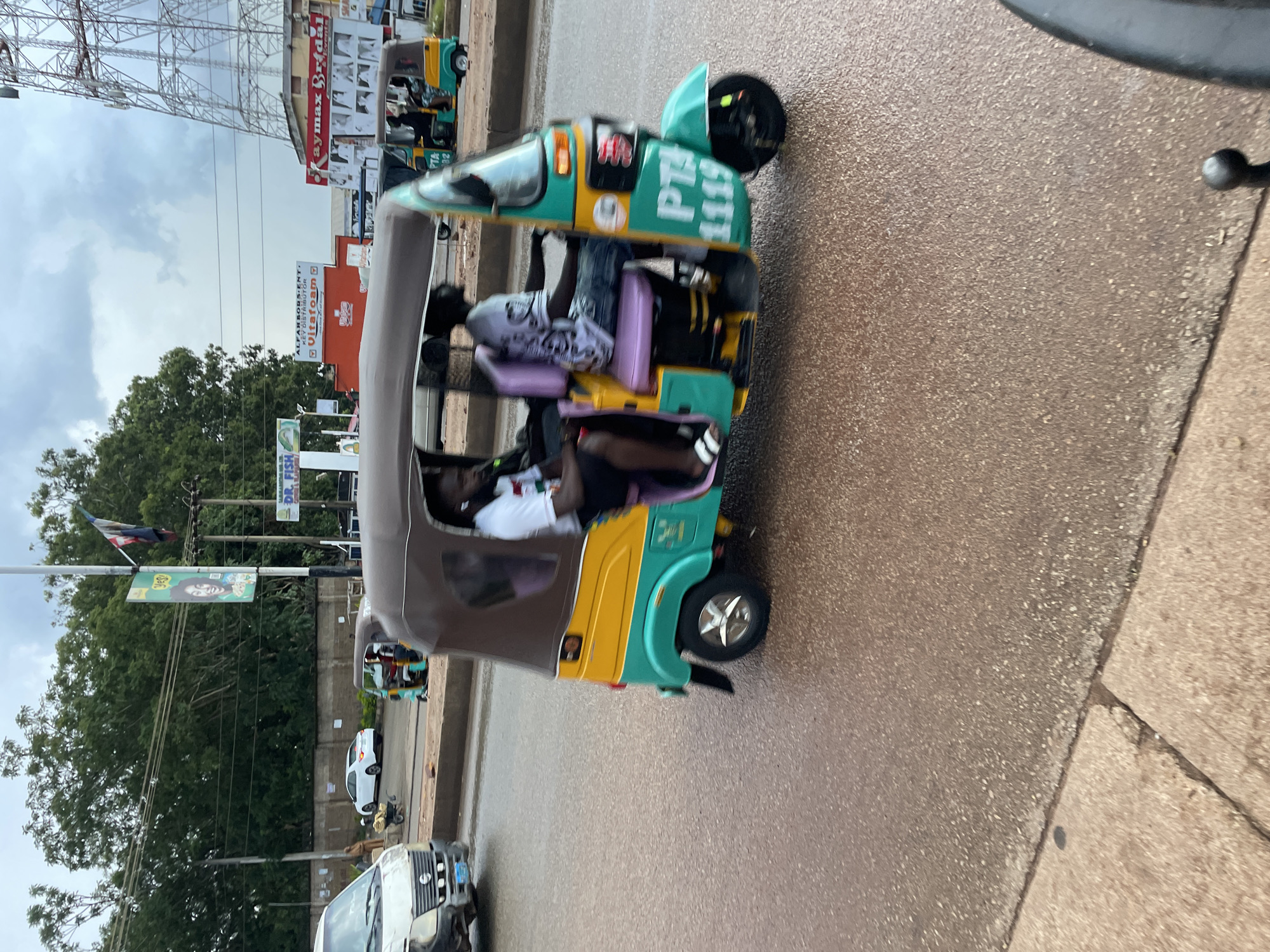 The image captures a street scene in Jos, Nigeria, under an overcast sky. In the foreground, a brightly colored auto-rickshaw, painted yellow and teal with a brown canopy, is in motion, its wheels blurred from movement. Two individuals are visible inside the rickshaw; one is driving, and another is seated behind him. The driver wears a white t-shirt with a graphic print and dark pants, while the passenger is in a white t-shirt and jeans. Both appear to be male.
The street surface is wet and appears to be asphalt. To the left of the rickshaw, a white van is partially visible, and further in the background, a white sedan is parked. A cart with some cargo is also visible.
The background features a lush green tree line, a tall cellular tower, and various buildings with signage. Prominent signs include "Kaymax Bridal," "Utam Dan," and a sign for "Dr. Fish." Utility poles with wires are also present. The overall impression is of a bustling urban environment.