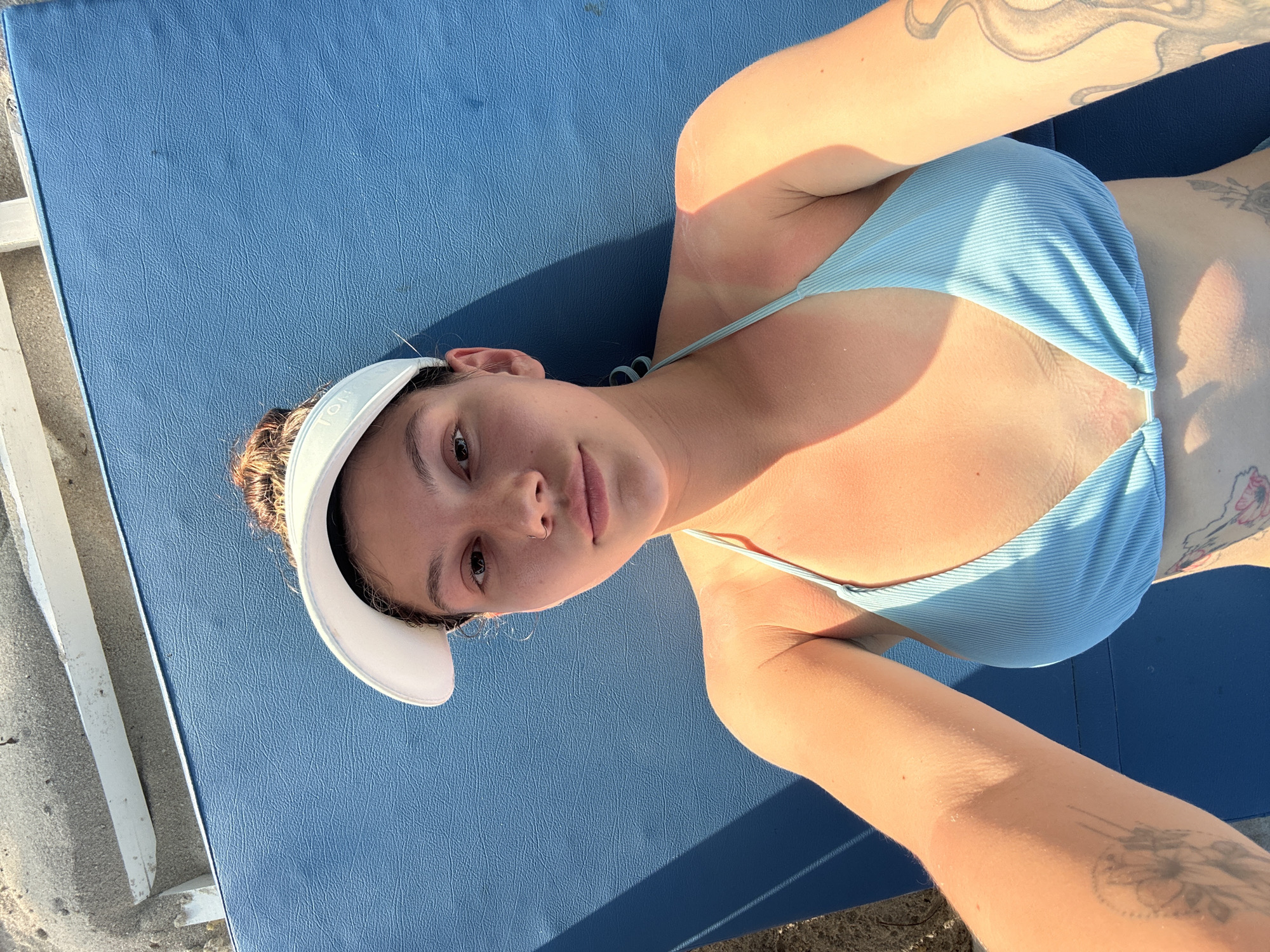 A young woman wearing a light blue bikini top and a white visor is lying on a blue cushioned surface. She is fair-skinned, has brown hair pulled back, and is facing the camera. She has tattoos on both arms. The scene appears to be outdoors, possibly at a beach or pool. The location is Baan Chaweng, Thailand.