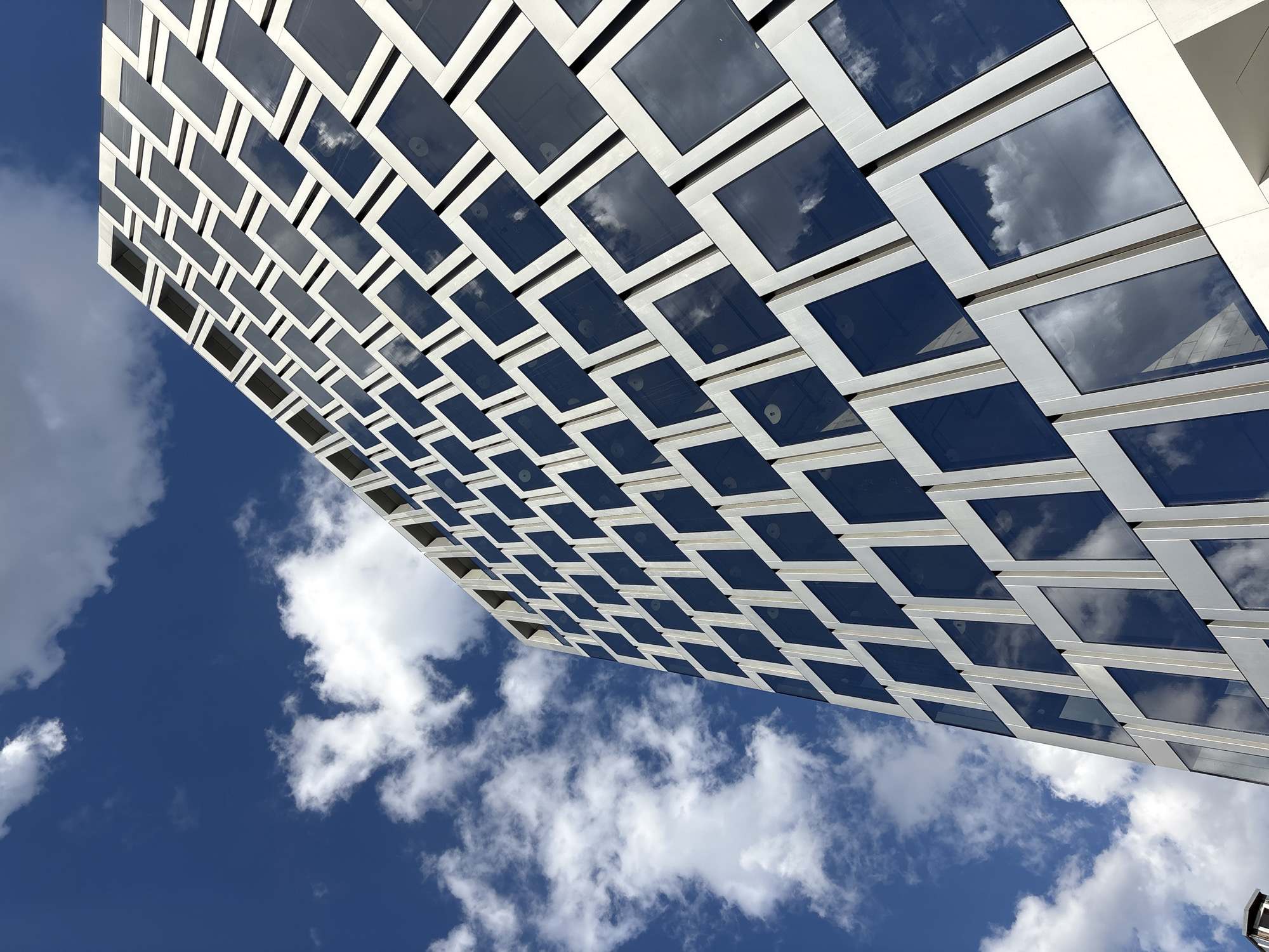 An extreme low-angle, wide shot captures the modern, glass-paneled facade of a building against a bright blue sky with white, scattered clouds. The building's design features a repeating pattern of rectangular dark glass panels framed by light-colored material, creating a geometric, textured appearance. The reflections of the sky and clouds are visible on the glass surfaces. The image is oriented vertically. The scene is devoid of people. The architecture suggests a contemporary urban setting, consistent with a modern office building or residential complex. The location is not explicitly indicated in the image, therefore its presence in Moscow, Russia cannot be confirmed.
