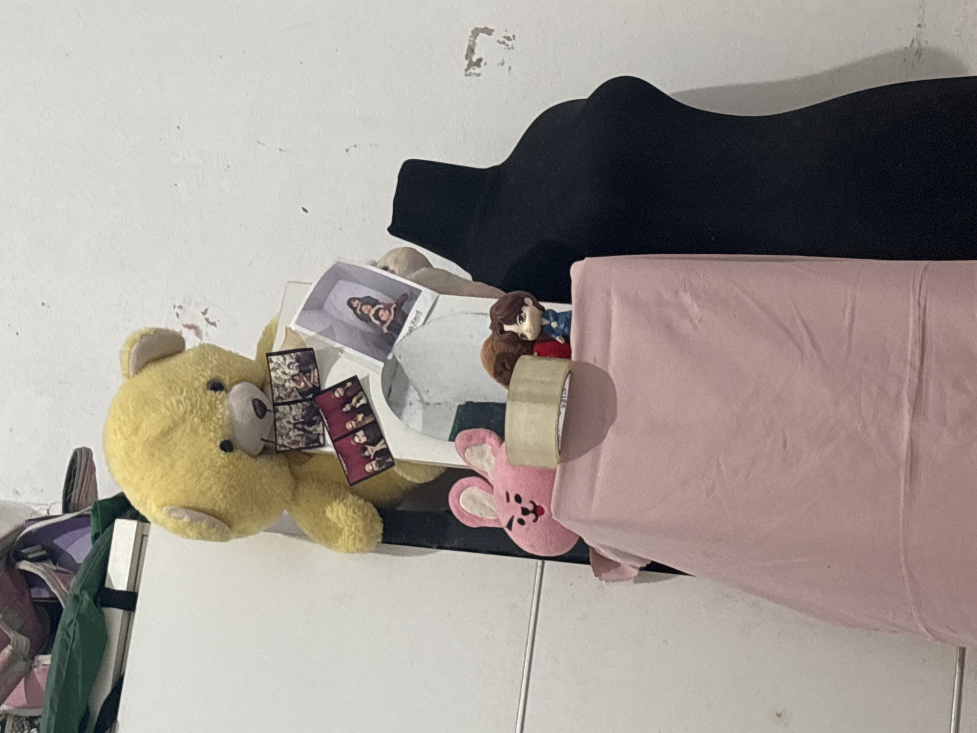 An indoor scene depicts a collection of decorative items against a textured white wall. A large, fluffy yellow teddy bear is positioned centrally, holding several small rectangular photographs. Below the teddy bear, to the left, is a pink plush bunny head with large ears and a black smiley face. Adjacent to the bunny head is a roll of transparent tape. To the right of the bunny head and tape is a miniature figurine of a male anime character. In the foreground, a large roll of light pink fabric is draped. Behind the fabric and figurines, a black, form-fitting garment, possibly a dress or a mannequin cover, is visible. The overall environment appears to be a domestic setting, potentially a room in North Jakarta, Indonesia, based on the provided context.