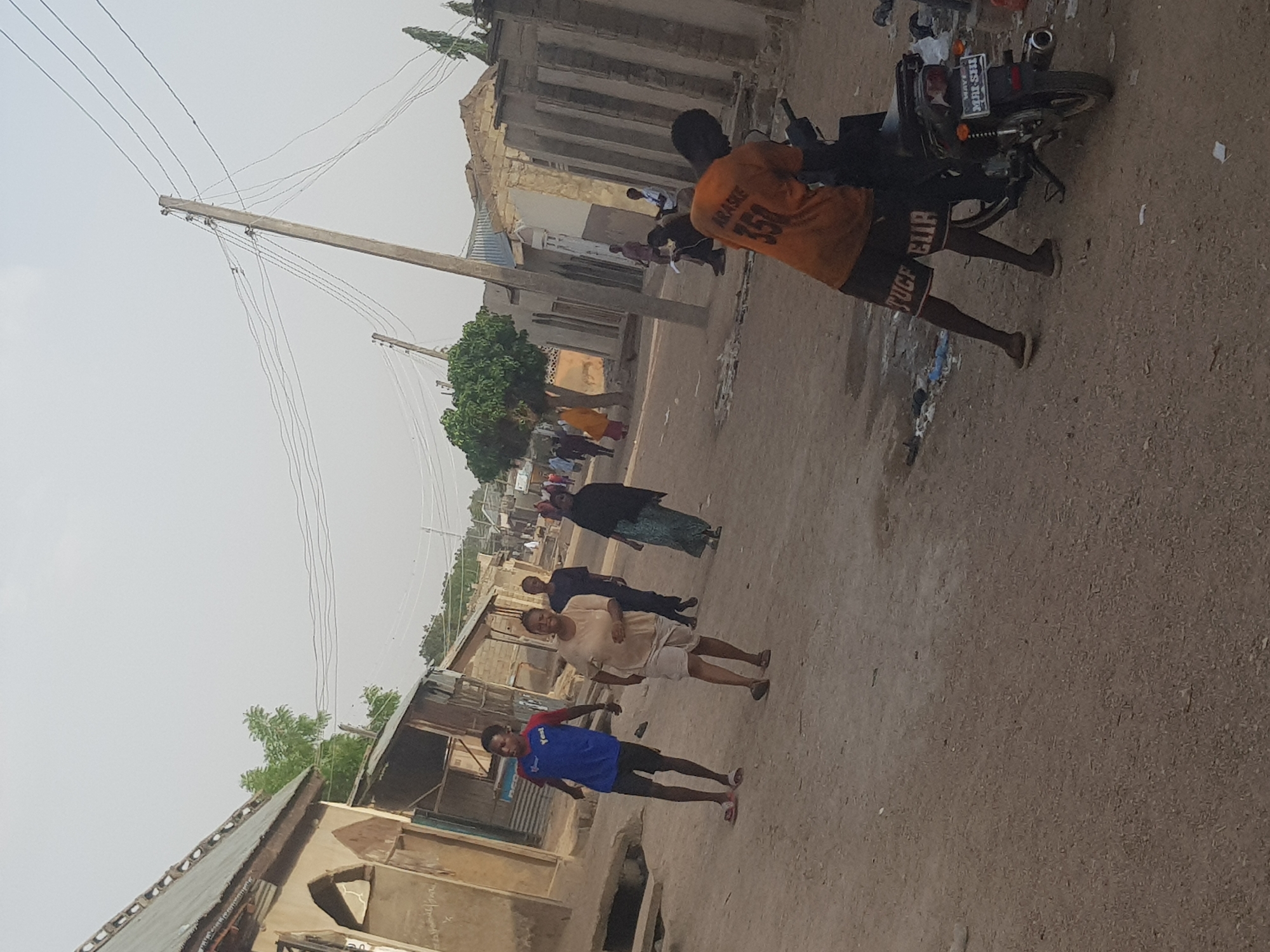 The image shows a street scene in Dutsinma B, Nigeria. A dirt road runs through the center, with buildings on either side. Several people are walking along the road. One man is standing next to a motorcycle. Power lines run along one side of the street. Trash is visible on the ground. The sky is overcast.