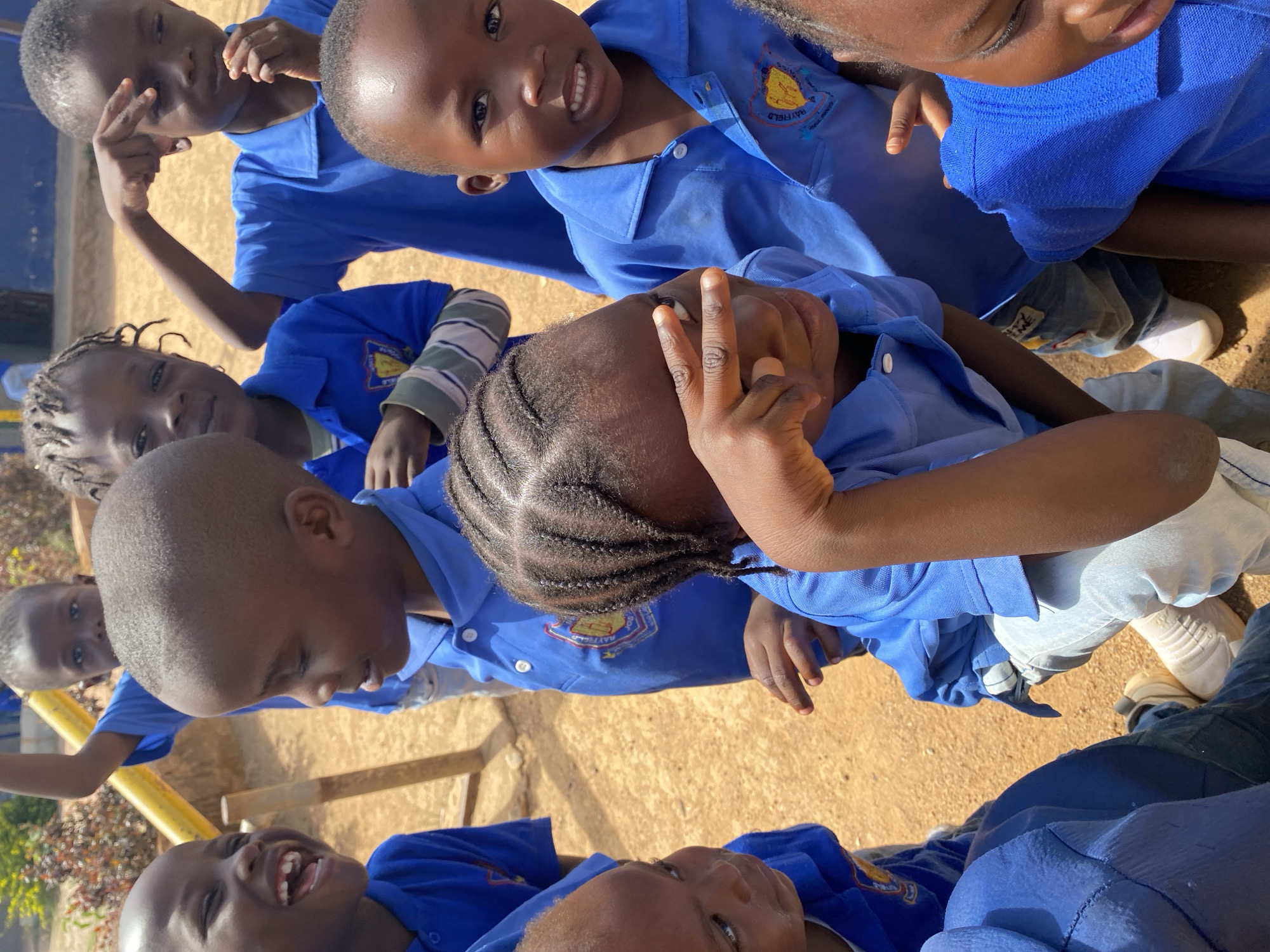 A group of children wearing blue school uniforms is gathered outdoors in Gura Top, Nigeria. The children are of African descent. The ground is dirt-colored. One child is flashing a peace sign. Some are smiling. A yellow metal structure is partially visible. Their uniforms have a Rayfield School emblem.