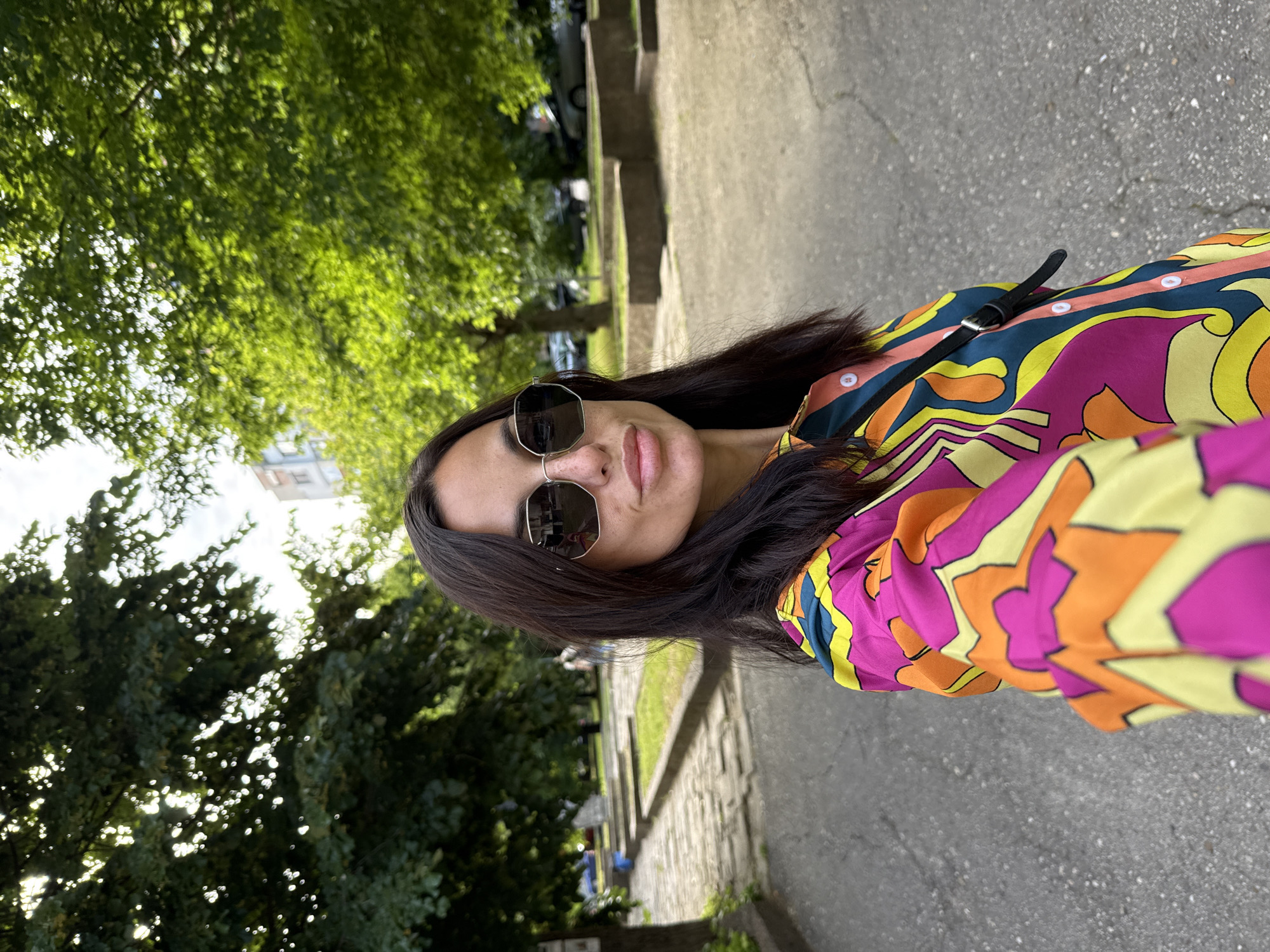 A woman wearing sunglasses and a colorful patterned shirt stands on a paved area outdoors. Behind her, trees with green leaves line a pathway leading up a slight incline. In the background, a multi-story building is visible. The scene appears to be a public space in the city of Pleven, Bulgaria, likely a park or residential area.