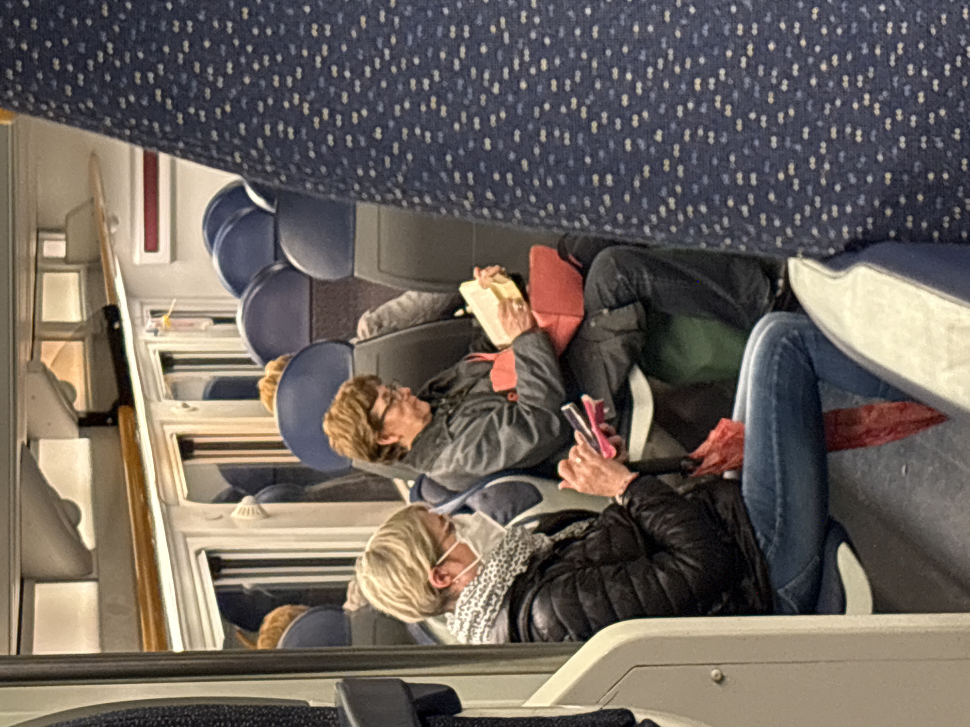 The image shows the interior of a train, likely in Milan, Italy based on the seating arrangement and interior design of the train. Three people are visible, all reclining in their seats. The person closest to the viewer is wearing a face mask, a black puffer jacket, and jeans. They are using a mobile phone. The person beside them is reading a book, wearing glasses and a jacket with red accents. The person behind them is partially visible. The seating upholstery is blue with a small dotted pattern. The window is to the left, showing reflections and hints of the outside world.
